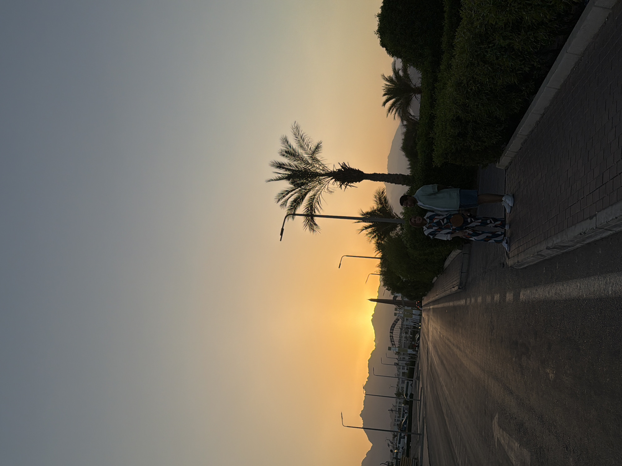 The image captures a sunset scene in Sharm El Sheikh, Egypt, looking west towards the mountains. Two people, a man and a woman, stand silhouetted on a paved walkway next to a road. The woman is wearing a patterned jumpsuit, and the man is wearing a light-colored short-sleeved shirt and shorts. They are facing each other and appear to be in conversation.
The sky displays a gradient of warm colors, transitioning from a bright orange near the horizon where the sun is setting behind the mountains, to a pale yellow, and then to a light blue at the top of the frame. Palm trees are visible on the right side of the image, with their silhouettes stark against the colorful sky. Streetlights are scattered along the roadside.
The road itself is paved and appears to be a main thoroughfare, with lane markings visible. Cars are present on the road in the distance, and buildings line the left side of the road, interspersed with more streetlights and some parked vehicles. The overall atmosphere is serene, typical of a dusk setting in a coastal resort town.