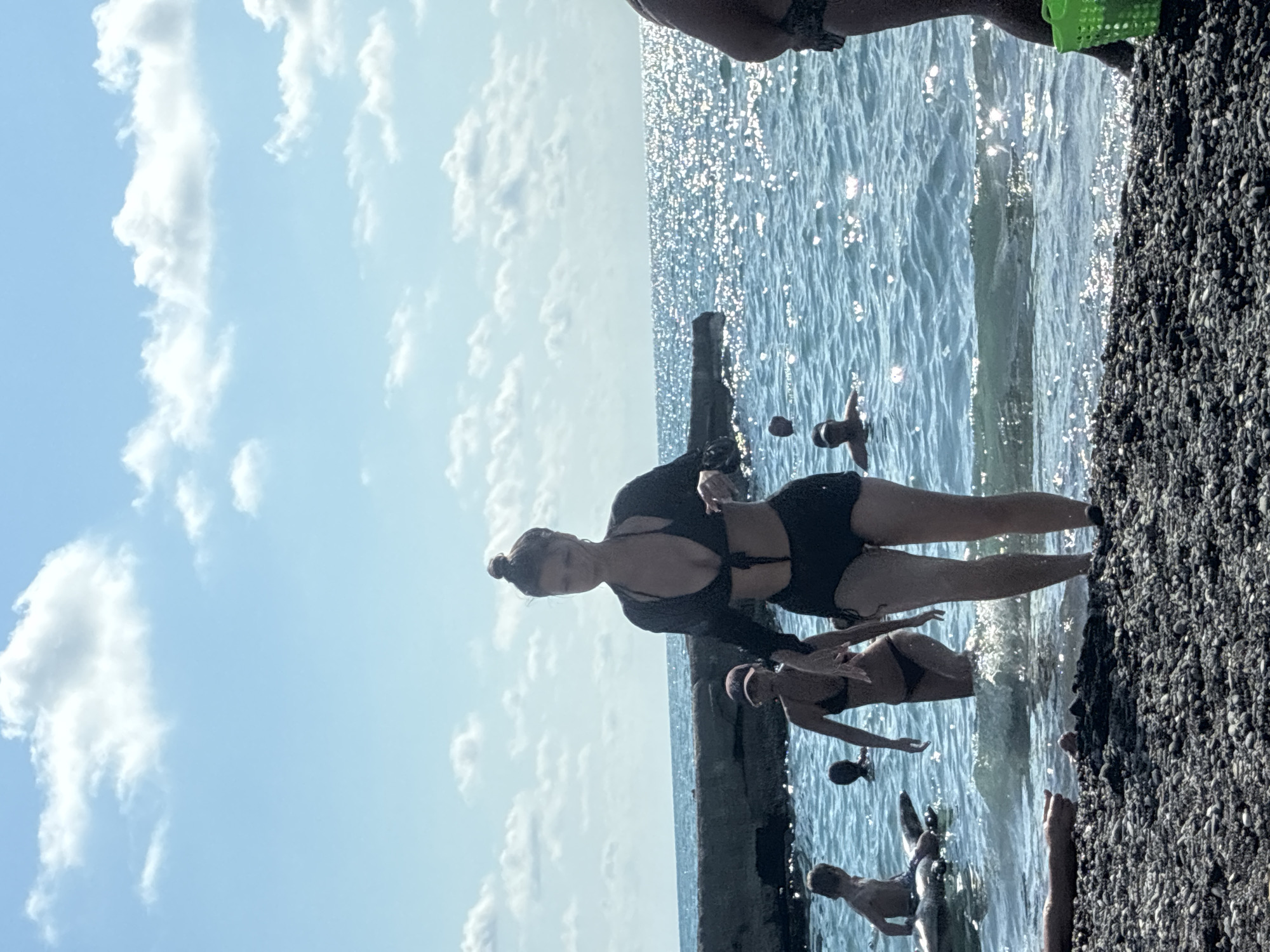 This media file captures a lively scene at the beach in Sochi, Russia. The primary subject is a woman in a black bikini, posing confidently by the water. She's the most prominent figure, her pose suggesting she's either posing for the camera or enjoying a moment of leisure.
Surrounding her are other individuals, though less distinctly visible, either in the water or on the shore. The water itself is a vibrant blue, reflecting the bright sunlight, indicating a clear, sunny day. The surface of the water is also dotted with glistening highlights, suggesting a slightly choppy but calm sea.
The setting appears to be a rocky beach, with dark, pebble-like stones making up the foreground on the right side of the image. A concrete structure, possibly a pier or a seawall, runs along the water's edge, providing a platform for the people in the scene.
The sky is a brilliant blue, adorned with fluffy white clouds, typical of a summer day. The overall atmosphere is one of recreation and enjoyment, a typical beach day experience. The time of day is clearly daytime, likely mid-morning to late afternoon, given the strong sunlight and the presence of people enjoying the beach. There are no visible text elements in the media file.