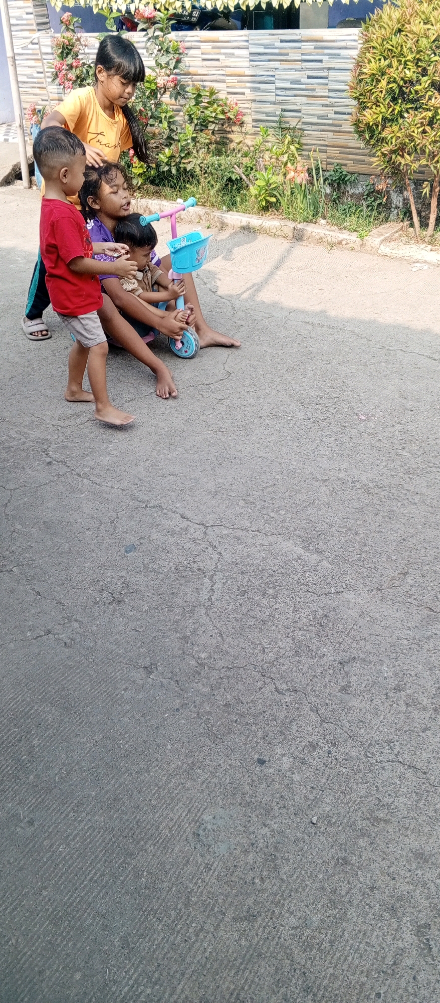 In Karawang, Indonesia, a group of four young children are gathered on a cracked asphalt surface. An older girl, wearing an orange t-shirt, stands to the left, with her hand resting on the head of another girl sitting down. The seated girl, in a purple shirt, is holding a blue plastic toy scooter with pink handlebars and a basket. A younger boy in a red t-shirt and light-colored shorts stands to the left of the seated girl, looking towards her. Another very young child, wearing a brown shirt, is being held by the seated girl. The background features a tiled wall, green bushes, and a small tree. The lighting suggests it is daytime.