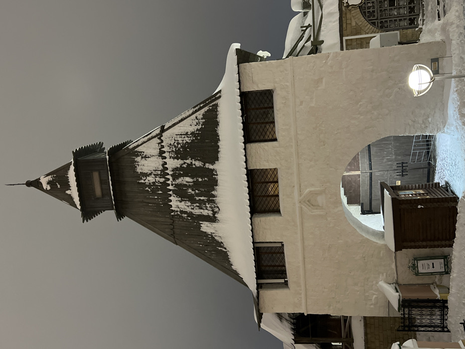 The image displays a snow-covered architectural structure at night, located in Matiushino, Russia. The central subject is a robust building, possibly a gatehouse, constructed with white, textured plaster walls and topped by a prominent, dark wooden conical roof. The roof features several tiers of dark wooden shingles, heavily blanketed with thick white snow along its eaves and sloped surfaces. The apex culminates in a smaller, belfry-like structure with a rectangular opening and a pointed finial.
Below the wooden roof, the white plastered facade contains three rectangular windows with dark frames and internal grid patterns, emitting a warm, artificial light from within. A large, rounded archway forms a passage through the base of the building at ground level. The ground, both leading to and within the archway, is covered in a substantial layer of pristine white snow.
To the left of the archway, a section of a black wrought-iron fence is partially visible amidst the snow. Adjacent to this fence stands a small, dark wooden kiosk or booth with horizontal slat siding, also partially obscured by snow. A white sign with dark text and a directional arrow is positioned near the kiosk. Inside the archway, a large double-leaf wooden gate with dark metal hardware is visible in the background, alongside a white metal barrier to its right. To the right of the archway, a modern street lamp featuring a spherical white diffuser on a dark pole illuminates the immediate foreground. Further right, a segment of an adjoining snow-covered building with an arched window is present. The sky above is uniformly dark, indicating nighttime conditions. No people, specific actions, or interactions are visible within the scene.