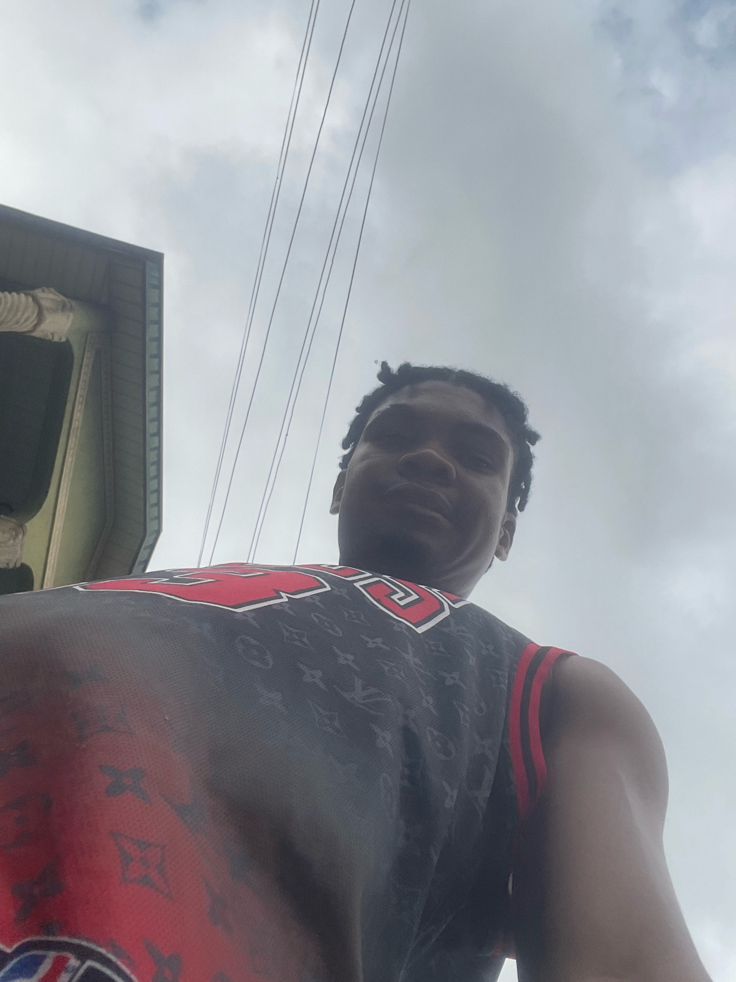 A low-angle, outdoor, medium shot captures a young Black man from the chest up, looking directly at the camera. He has dark, braided hair and is wearing a black jersey with a repeating Louis Vuitton pattern in a darker shade. Red trim accents the edges of the jersey, including stripes on the shoulder. Behind him, a cloudy, overcast sky dominates the background, with several utility wires running horizontally across the frame. To the left of the man, a portion of a building is visible, with a light-colored decorative element on its exterior and a green-painted section. The scene appears to be set in a city environment.