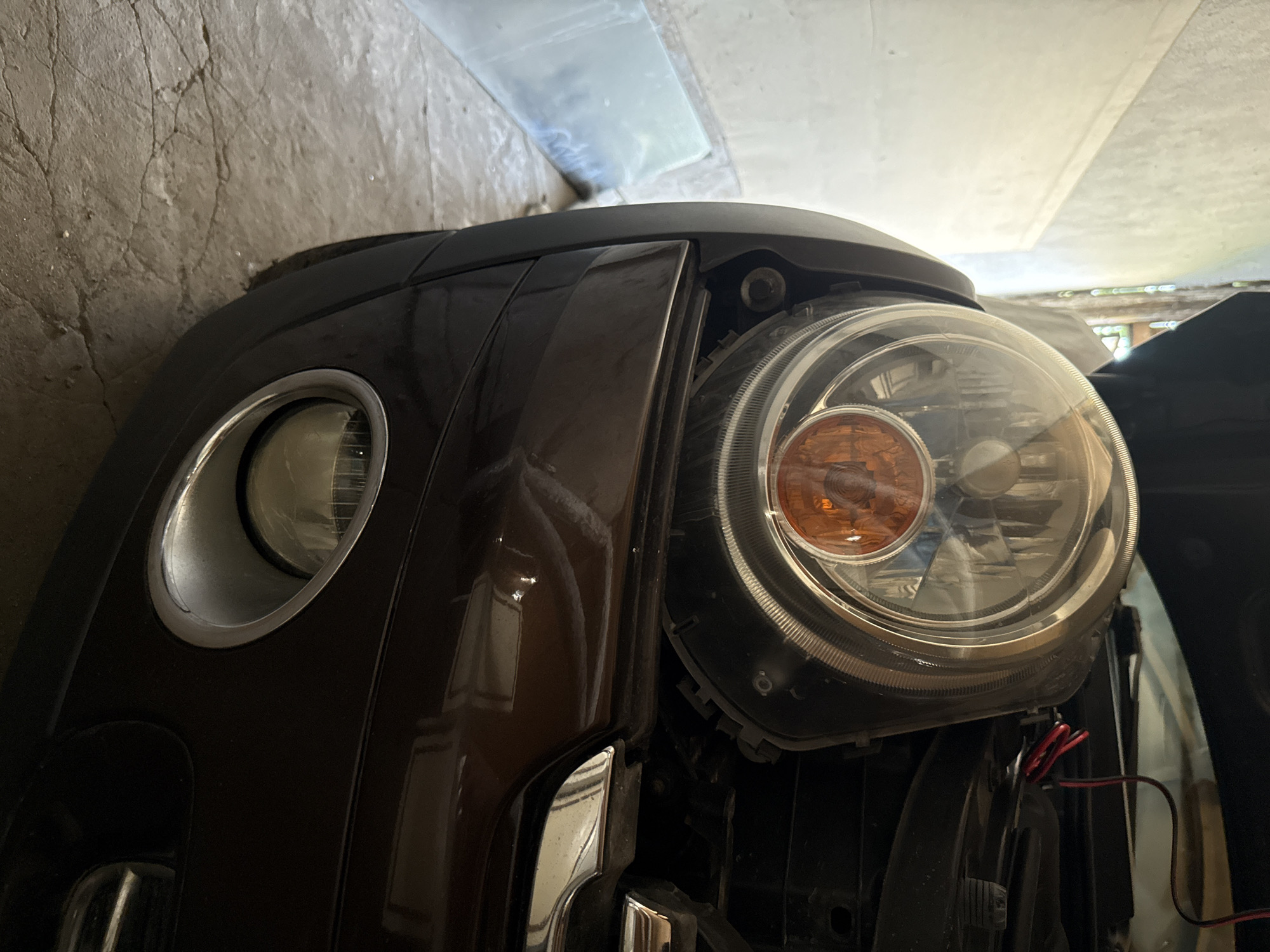 A close-up, eye-level, medium shot captures the front left side of a dark brown Mini Cooper car. The car's hood is open, revealing the headlight assembly and surrounding engine components. The headlight is large and round with a clear lens, exposing the orange turn signal bulb and various reflectors within. The bumper is visible below the headlight, featuring a circular fog light with a chrome bezel. The car is parked on a cracked concrete floor, possibly in a garage or workshop, indicated by the wall in the background. The environment is dimly lit with natural light filtering in from an unseen source. No people or animals are present.