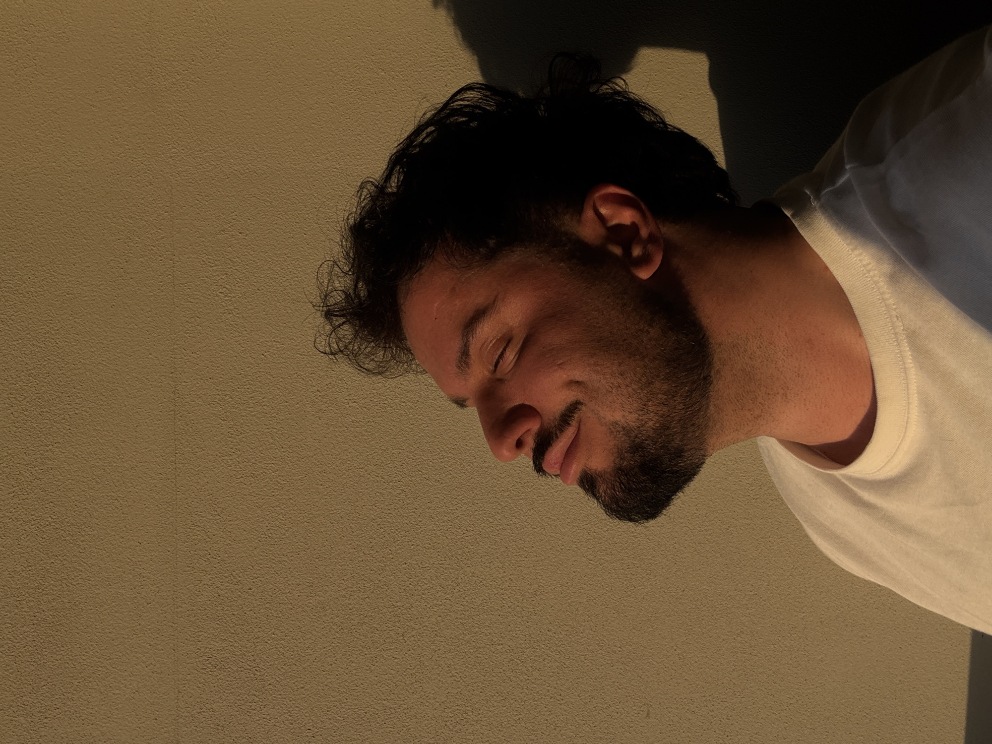 The image presents a rotated, close-up portrait of a man, likely in Milan, Italy. He is depicted against a textured, light brown wall, possibly lying down or leaning. The lighting is warm and golden, characteristic of sunset or golden hour, casting a distinct dark shadow behind his head. The man has dark, curly hair and a neatly trimmed beard. He is wearing a plain white t-shirt. His eyes are closed or squinted, and a subtle, contented smile or smirk plays on his lips, conveying a sense of peace or relaxation. There is no visible text in the image.