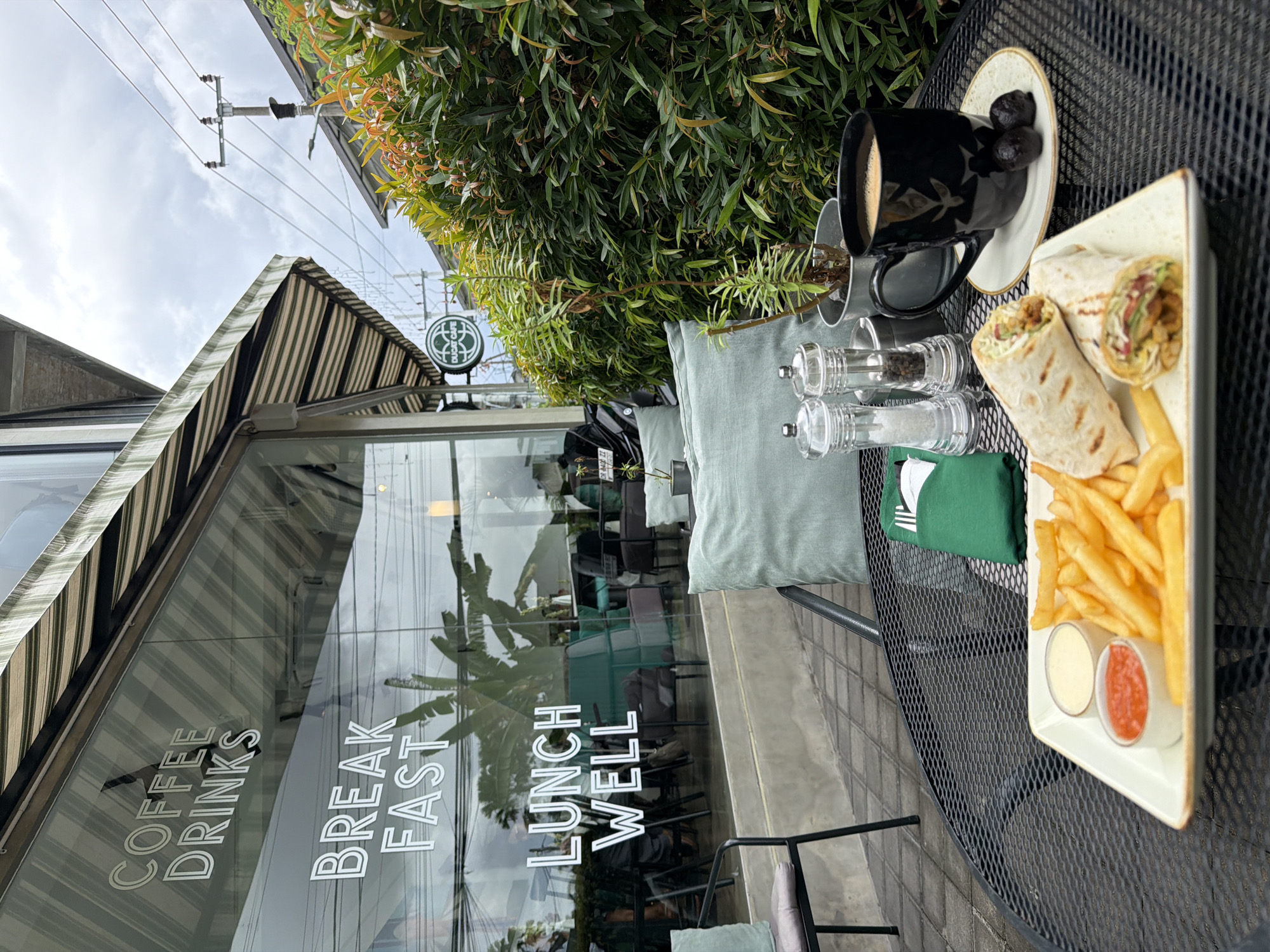 The image depicts a table setting outside the Ducat Cafe in North Kuta, Indonesia. The table, made of black metal mesh, holds a plate with a wrap, fries, and two dipping sauces, alongside a cup of coffee with dates, salt and pepper shakers, a green napkin, and a cushion. The cafe's window displays the words "Coffee Drinks," "Breakfast," and "Lunch Well." A green sign with the cafe's name is visible above the window. A lush green bush is positioned behind the table. Overhead electrical wires are visible against a cloudy sky. The cafe is reflected in the window, showing chairs and plants inside.