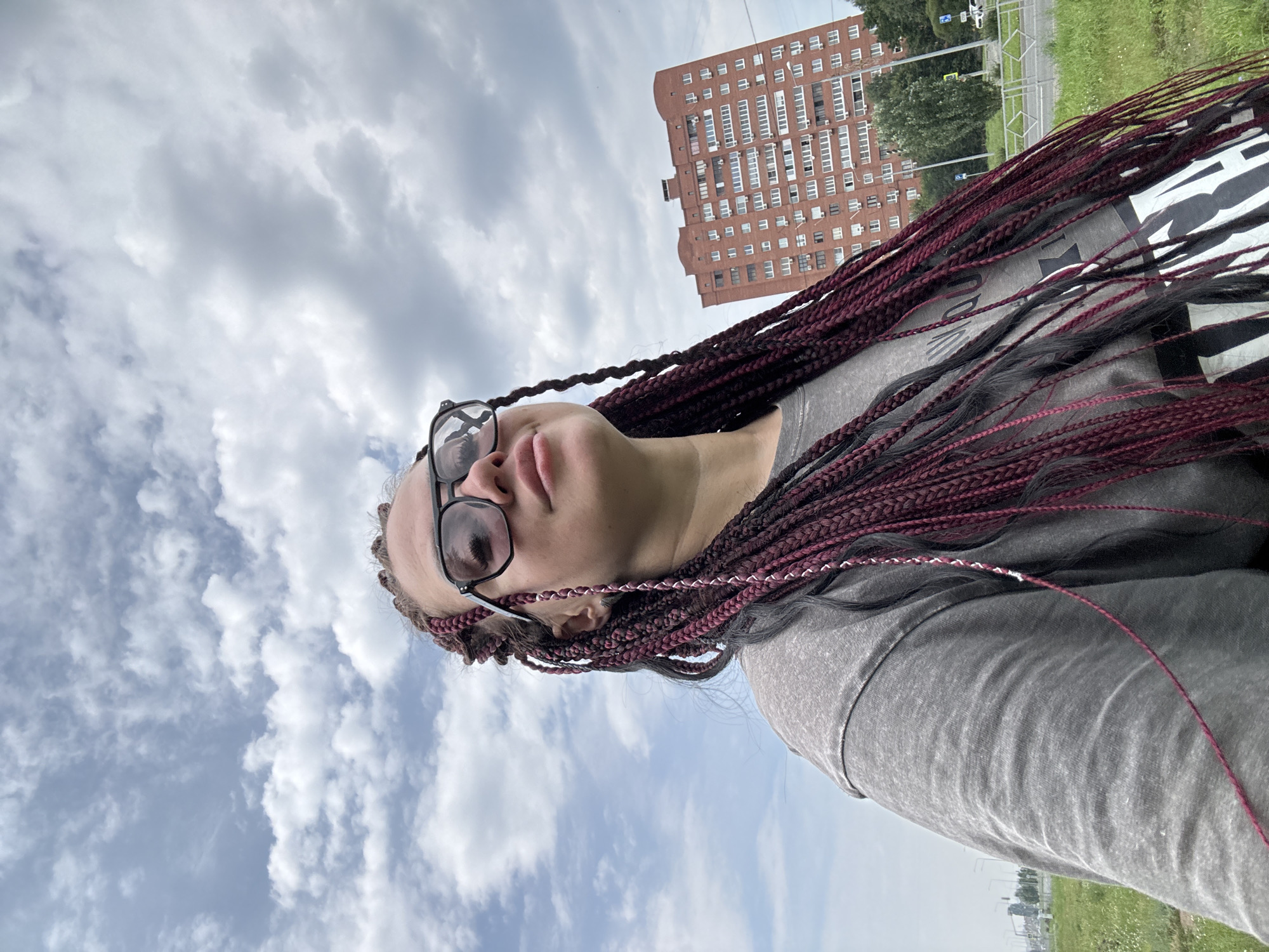 A young woman with burgundy and black box braids and large black-rimmed glasses is photographed from a low angle looking up at a cloudy sky. She is wearing a grey t-shirt. In the background, a tall, multi-story brick apartment building with many windows is visible, along with green grass and some trees. The setting appears to be an outdoor urban environment.