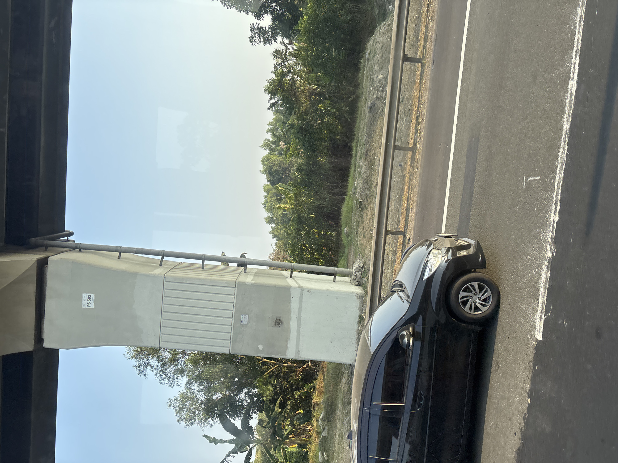 This image captures a scene on the outskirts of Pasirtanjung, Indonesia. A black car is traveling on a paved road, indicated by the presence of lane markings. To the left of the road, a large concrete pillar, part of an elevated structure, stands tall against a clear blue sky. This pillar has a small white sticker on its surface with the text "PS 502". Beyond the pillar, dense green vegetation and trees suggest a rural or semi-rural environment. The overall lighting indicates it is daytime, likely morning or afternoon due to the clear sky and shadows.