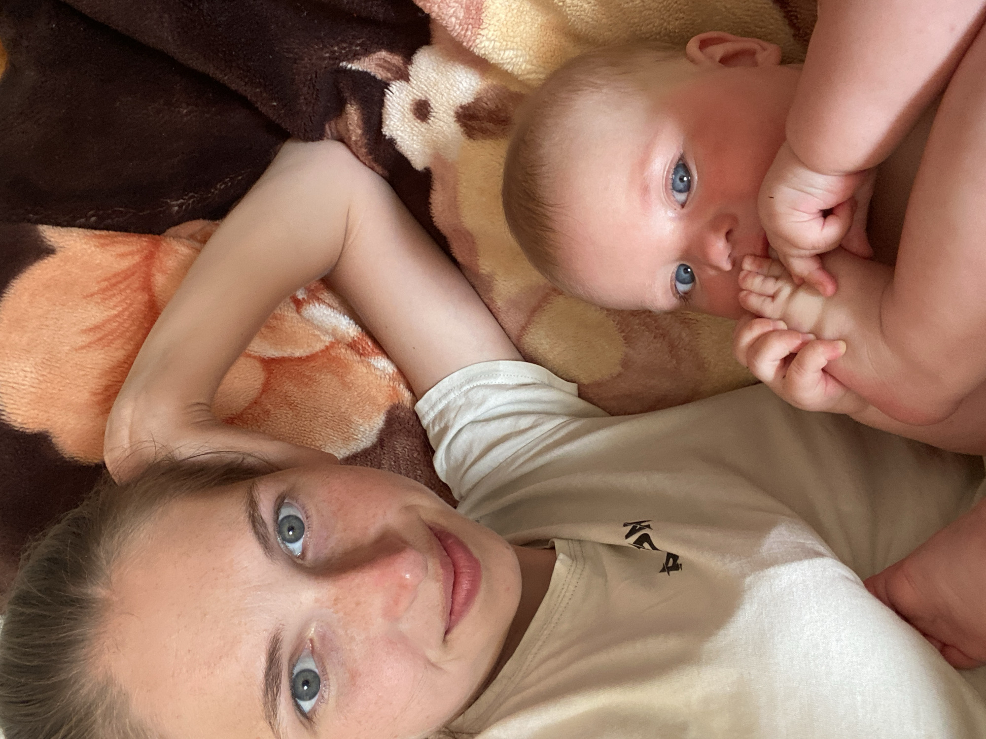 The media file is a close-up, overhead shot of a woman and a baby lying on a blanket. The woman is in the foreground, her face is turned towards the camera, and she is smiling. Her eyes are wide open, and she has light-colored hair pulled back from her face. She is wearing a light-colored t-shirt.
The baby is positioned in the upper right portion of the frame, lying on its back. The baby has bright blue eyes and is looking up at the camera. One of the baby's hands is near its mouth, and the other is resting on its chest. The baby's skin is fair, and it appears to be wearing a light-colored garment.
They are both lying on a soft blanket with a floral pattern. The blanket has brown, orange, and beige tones, with some lighter cream-colored areas. The lighting suggests it is daytime, and the setting appears to be indoors, possibly a bedroom or living room, given the presence of the blanket. The overall mood is warm and intimate, with both subjects looking relaxed and happy. There is no visible text in the media file.