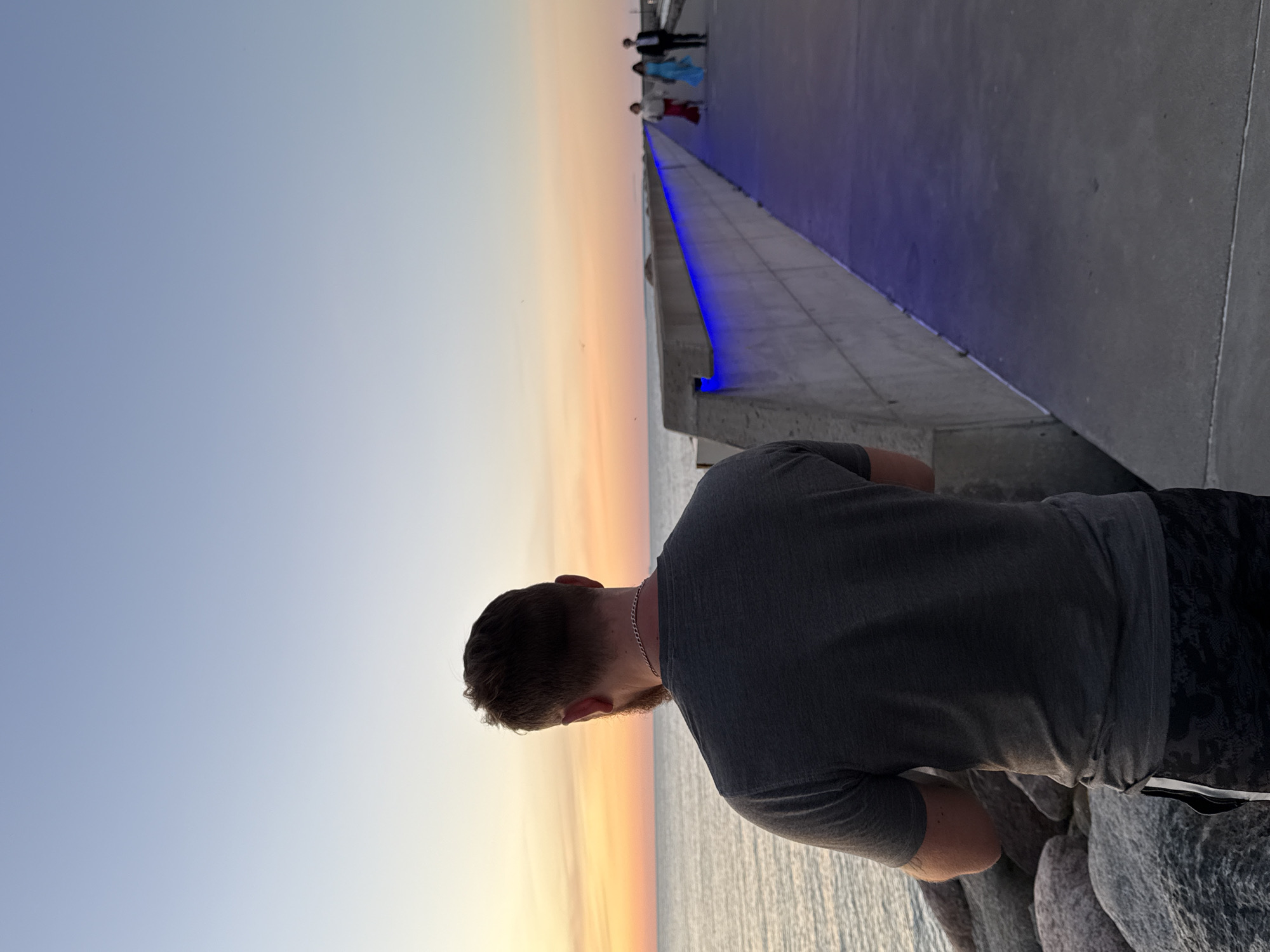 A man stands facing away from the camera, looking out at a body of water during sunset. He is wearing a dark gray t-shirt and black patterned shorts. To his right, a concrete walkway extends into the distance, illuminated by blue lights embedded in the wall. Three people, two women and a man, are walking on the walkway further down. The sky is a soft gradient of blue and orange, typical of a sunset. The scene appears to be a coastal area, possibly a pier or a breakwater. The man is standing near a pile of large rocks on the left side of the frame. The location is identified as Tallinn, Estonia.