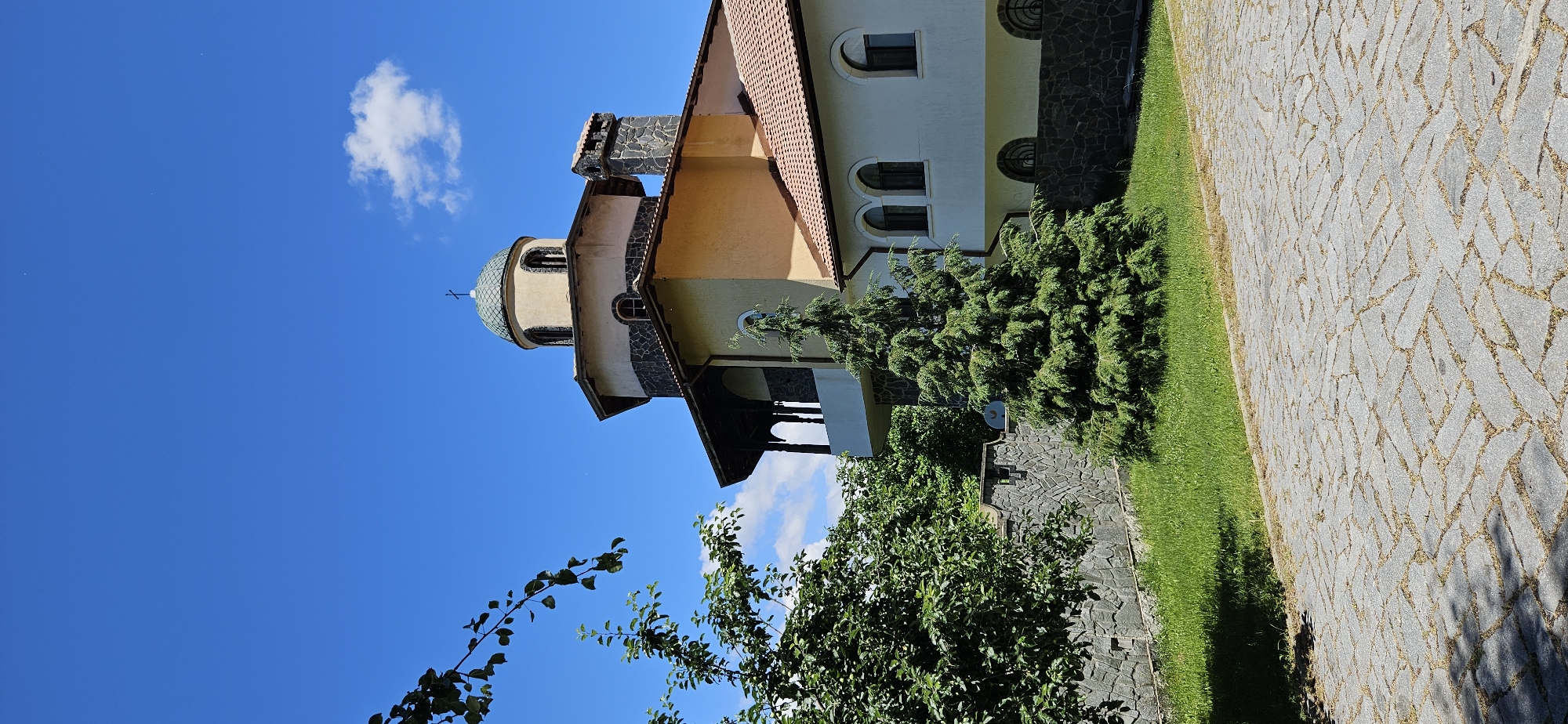 The image depicts a bright, sunny day at a building with architectural features resembling a church or monastery, located in Ovchartsi, Bulgaria. The building has a light yellow facade, a domed roof topped with a cross, and an octagonal section with dark stone accents. A chimney made of stone is visible on one side of the roof. The building is surrounded by greenery, including a tall, conical evergreen tree and lush grass. A stone wall separates the grassy area from a paved pathway made of irregular stone blocks. The sky is a clear, vibrant blue with a few scattered white clouds. A tree branch with green leaves is in the foreground on the left side of the image. The scene appears peaceful and serene.