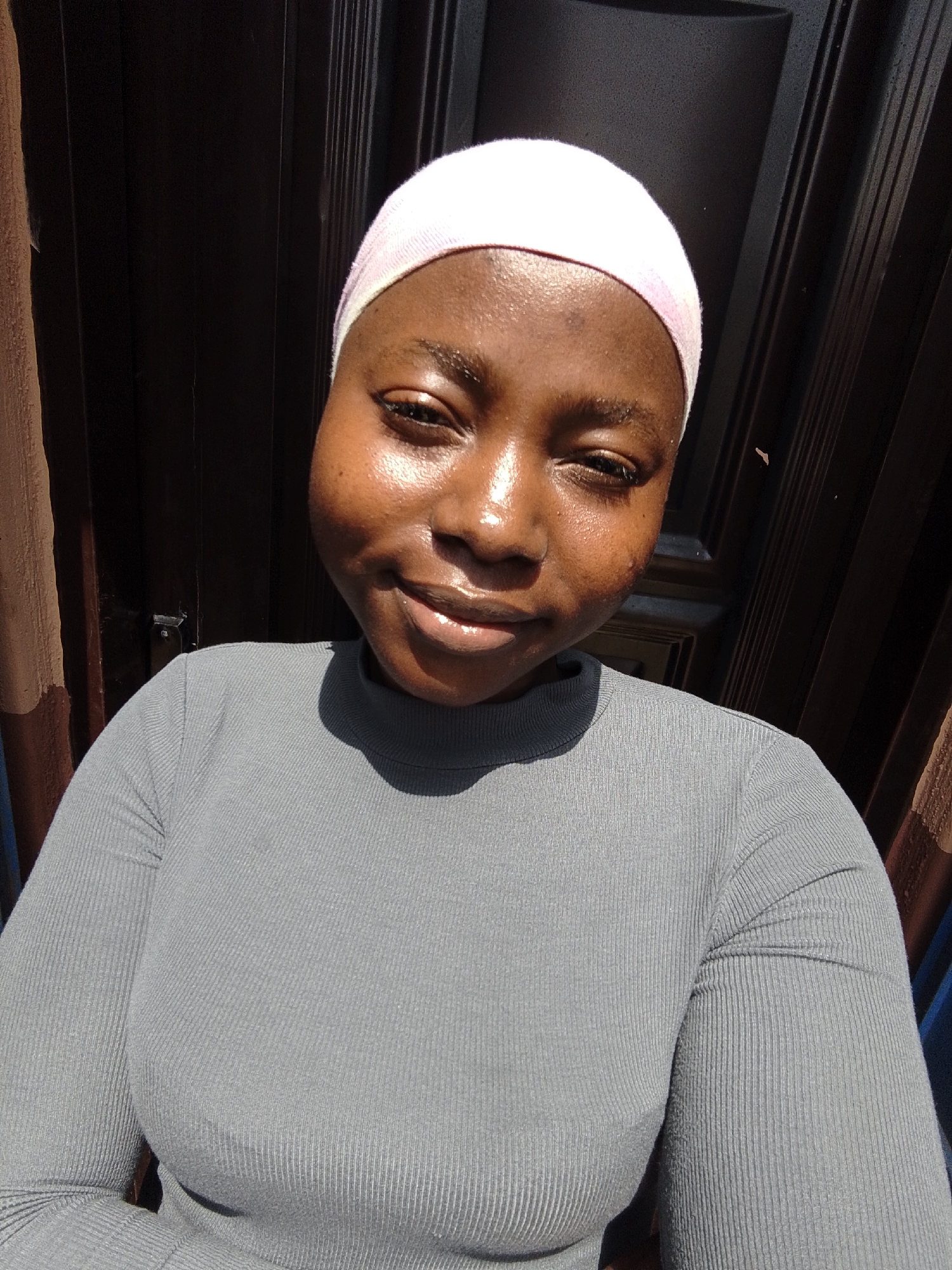 A young Black woman is pictured in a close-up selfie, looking directly at the camera with a slight smile. She is wearing a light pink headscarf and a grey, ribbed long-sleeved shirt. The lighting suggests it is daytime, with sunlight illuminating her face, creating highlights and shadows. The background is dark and textured, appearing to be a doorway or a wall with vertical panels. To the left of the frame, a curtain or fabric of a light brown hue is visible. The woman's skin is smooth and appears to have a dewy sheen, possibly due to sweat or a cosmetic product. Her eyes are dark and her lips are slightly parted, also appearing glossy. The overall mood is relaxed and casual. No text is visible in the image. The image was taken in Jos, Nigeria.
