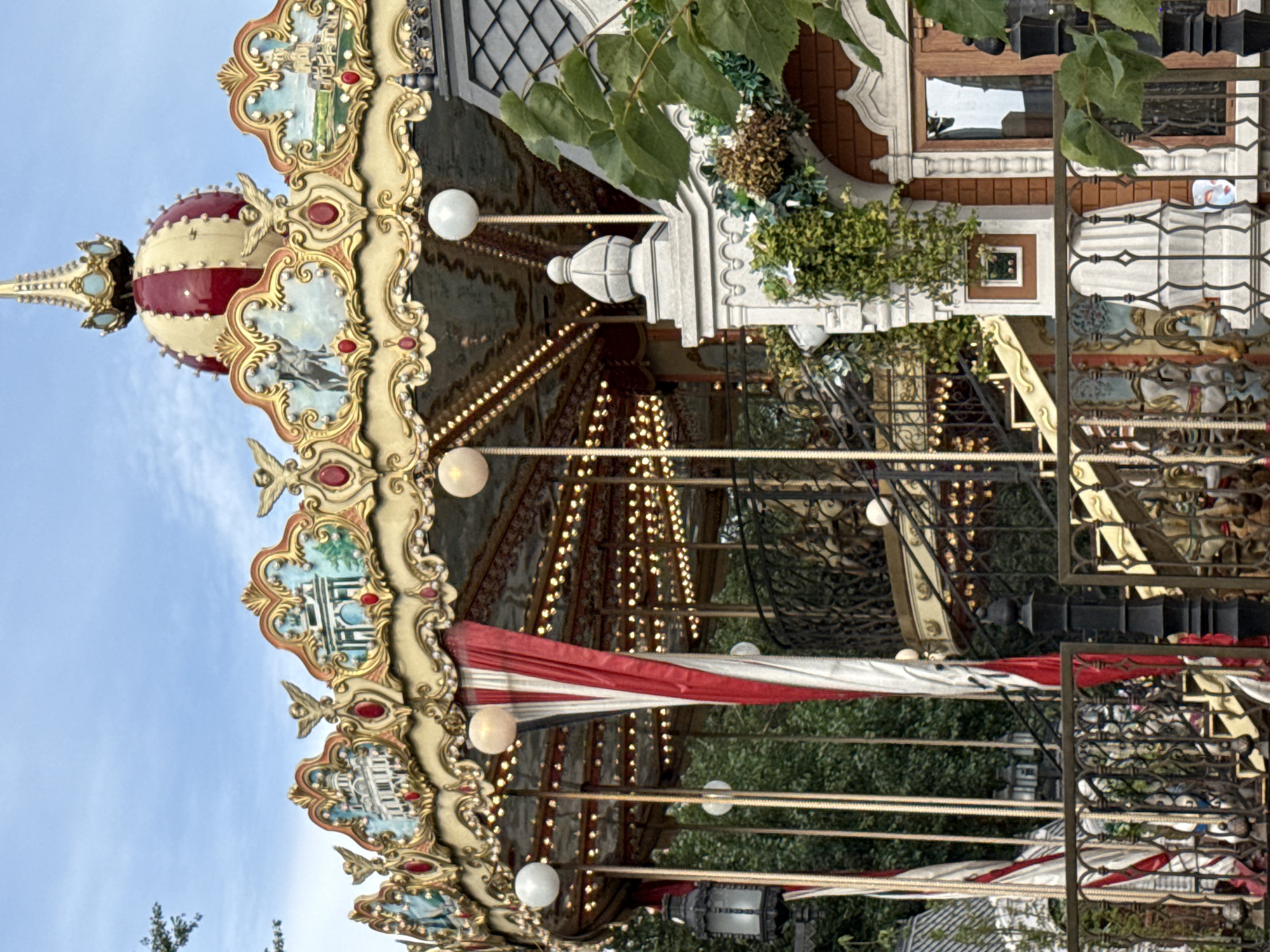 The image shows a detailed view of a carousel, likely a European-style carousel, given its ornate design. The carousel features a brightly colored canopy with elaborate gold trim, adorned with painted scenes of buildings and cherubic figures. Red and white striped fabric hangs between the poles supporting the canopy, and a string of lights is visible around the edge. At the very top, a domed structure with red and gold accents serves as the carousel's pinnacle.
The carousel is situated outdoors, with lush green trees visible in the background, suggesting a park or amusement area. A section of a building with a grey tiled roof and brick facade is visible on the right side of the image, partially obscuring the carousel. This building has a wooden-framed window and is decorated with ivy and other greenery. A black metal fence with intricate scrollwork is in the foreground, further framing the scene. The overall impression is one of festive grandeur, characteristic of a classic amusement ride. The style and design of the carousel, especially the painted murals and gilded ornamentation, suggest it might be a historically inspired or vintage piece.