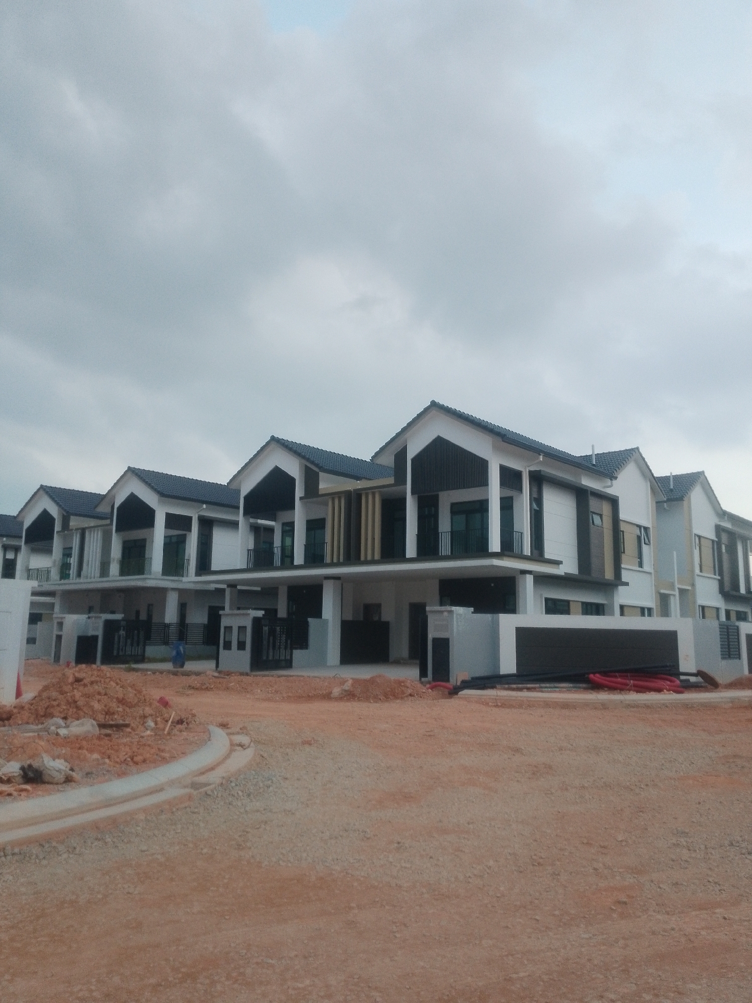 The image depicts a row of modern, newly constructed houses in a development project located in Iskandar Puteri, Malaysia. The houses are two stories high with white and grey facades, dark tiled roofs, and dark-colored window frames and balconies. Some houses feature decorative vertical wooden slats. The foreground shows a construction site with dirt, gravel, and unfinished concrete pathways and curbs. Piles of dirt and construction materials, including red and black pipes, are visible. The sky is overcast and cloudy. A single person, wearing blue clothing, is visible walking in the distance near the houses. The scene suggests an active construction phase of a residential neighborhood.