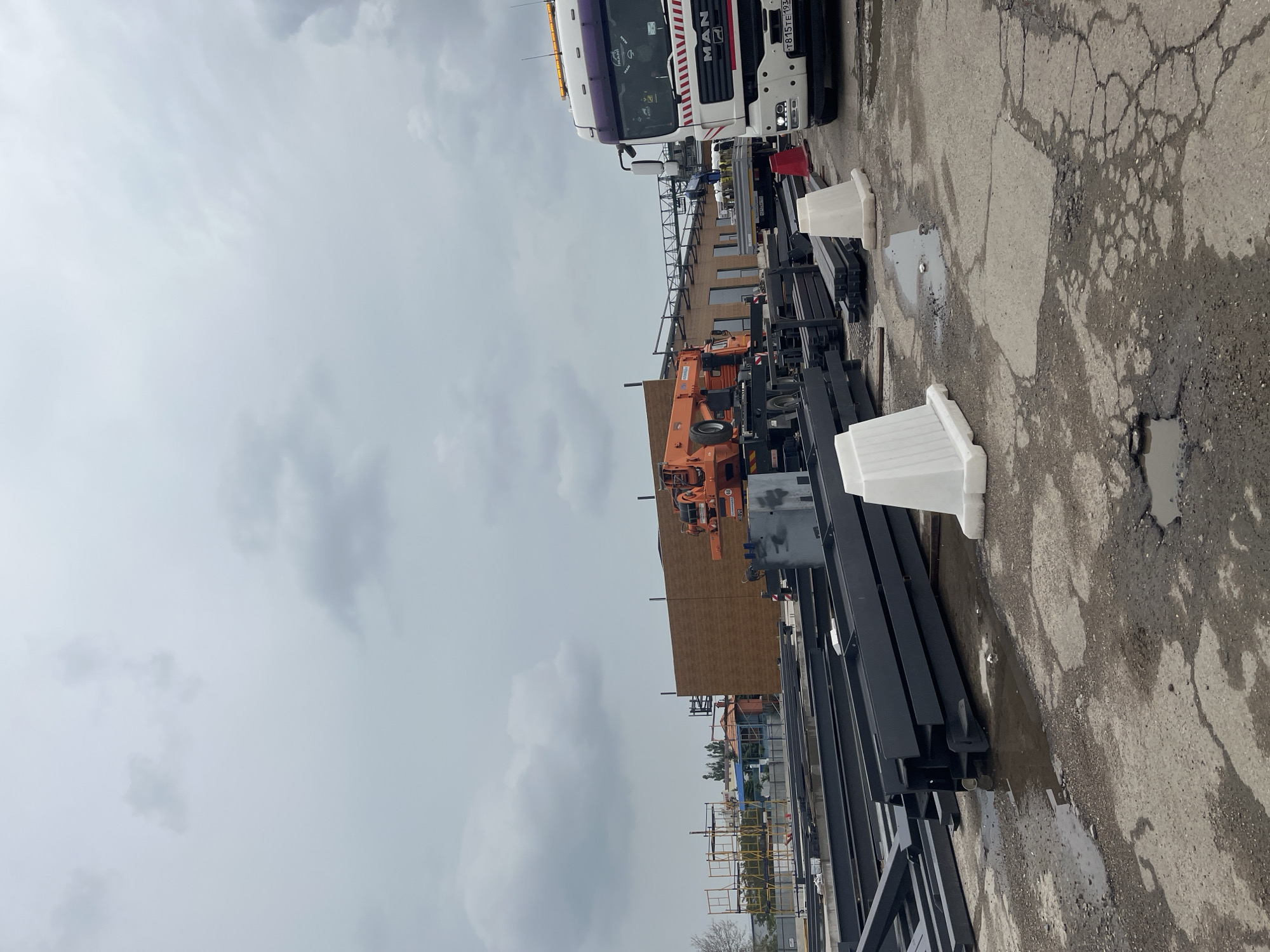 The image depicts an outdoor scene likely associated with construction or industrial activity. The sky is overcast. A MAN truck is parked along the right edge of the image. There is construction equipment, including an orange crane, a multi-level metal structure, and stacks of black metal beams laid on the cracked concrete ground. White plastic barriers are positioned around the equipment. There are puddles of water on the ground, suggesting recent rainfall. Scaffolding is visible in the background. Given the presence of a truck with Russian license plates and other details, the location is possibly a construction or industrial site in Krasnodar, Russia. There are no people visible in the image.