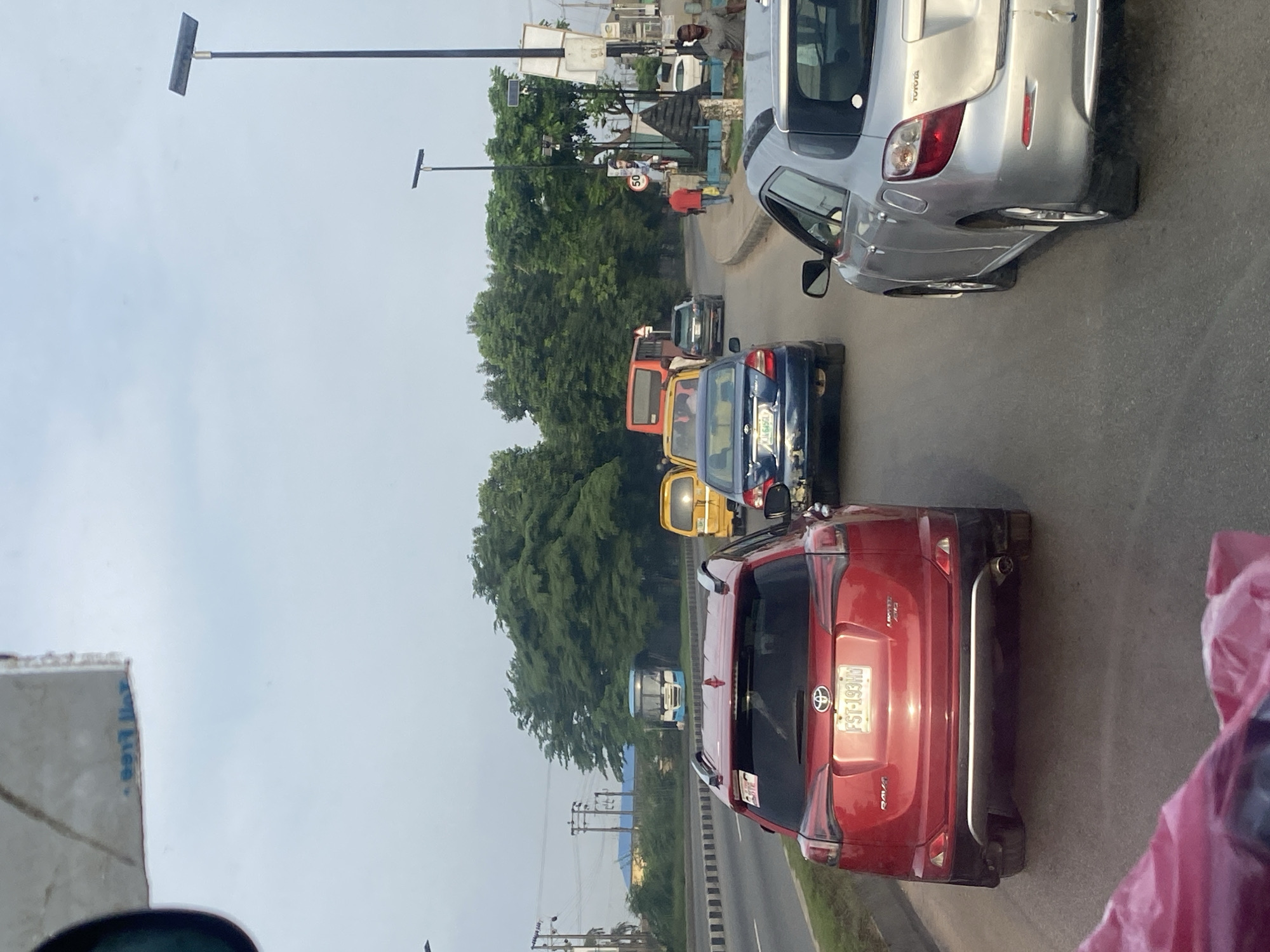 The image captures a traffic scene in Ikorodu, Nigeria, under an overcast sky. The foreground is dominated by the rear of a red SUV, likely a Toyota RAV4 judging by the badging, with the license plate FST-193HY visible. To the right of the red SUV is the rear of a silver Toyota hatchback. In front of these vehicles, a blue sedan is visible, followed by a yellow minivan or bus. Further ahead, an orange bus is seen in traffic. The road appears to be a multi-lane highway with trees lining the median and the background. Streetlights are present along the road. People are visible on the sidewalks and near some of the vehicles. The overall impression is one of moderate traffic congestion on a main thoroughfare in Ikorodu.