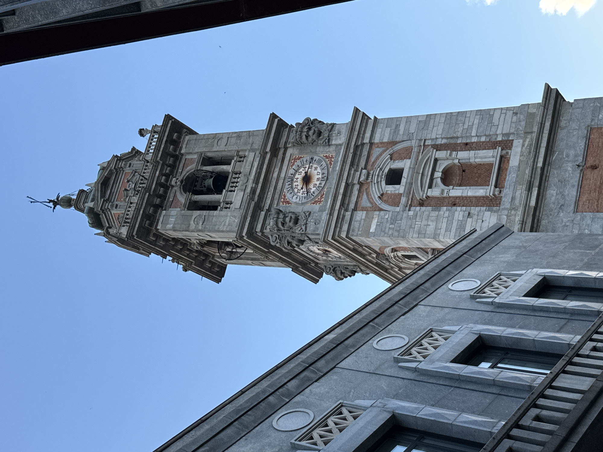The image shows the clock tower of the Basilica di San Vittore in Varese, Italy, against a clear blue sky. The tower is made of stone and brick, with a large clock face featuring Roman numerals and a 24-hour dial. Decorative carvings of lion heads adorn the tower. To the left, a modern building with large windows and a grey stone facade is visible. The perspective is from a low angle, looking up at the tower and the building.