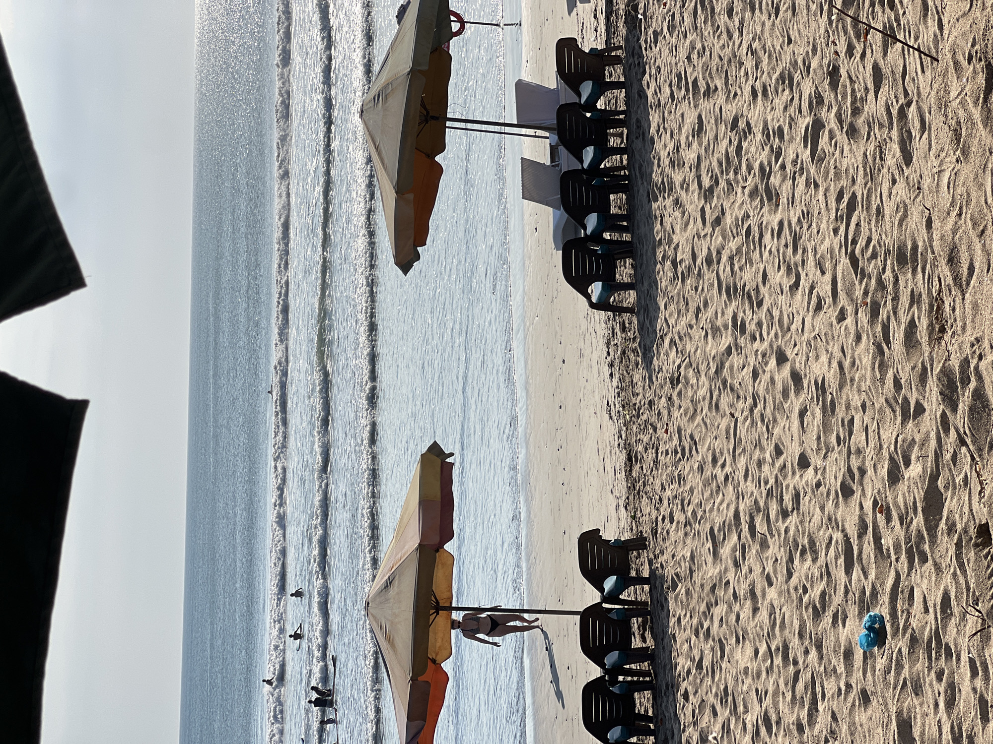 This is a medium-shot image of Kuta Beach in Kuta, Indonesia, on a sunny day. The foreground is dominated by textured sand with footprints and a small blue object. In the mid-ground, two beach umbrellas provide shade for rows of empty lounge chairs. On the left, a woman in a bikini walks away from the camera toward the ocean. The ocean is calm with gentle waves and several surfers are visible in the water. The horizon line is clear, and the sky is bright with a slight haze.