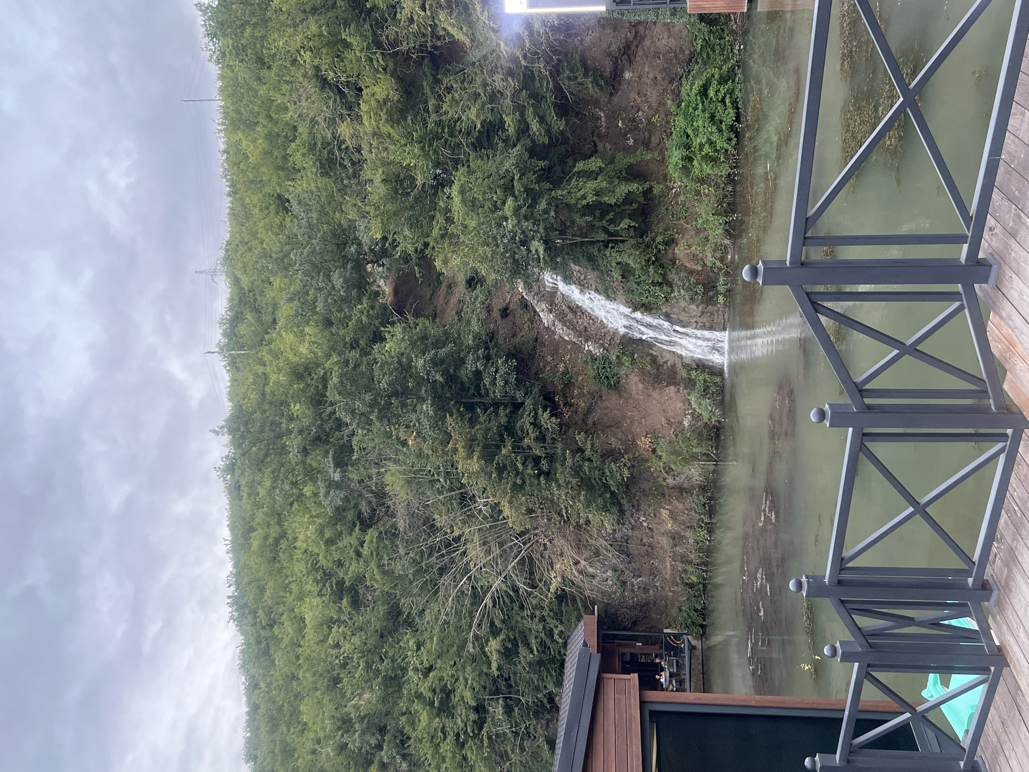 A waterfall cascades down a wooded hillside into a placid body of water. A modern wooden building with a dark roof is situated on the left side of the frame, near the water's edge. In the foreground, a dark metal railing with a criss-cross design separates a wooden deck from the water. The water appears somewhat murky and contains scattered debris. The hillside is densely covered with trees and vegetation, and the sky is overcast. In the distance, utility poles and power lines are visible atop the hill. Two individuals are seated at a table on a covered porch area of the building. The scene evokes a natural, serene setting with man-made structures integrated into the landscape.