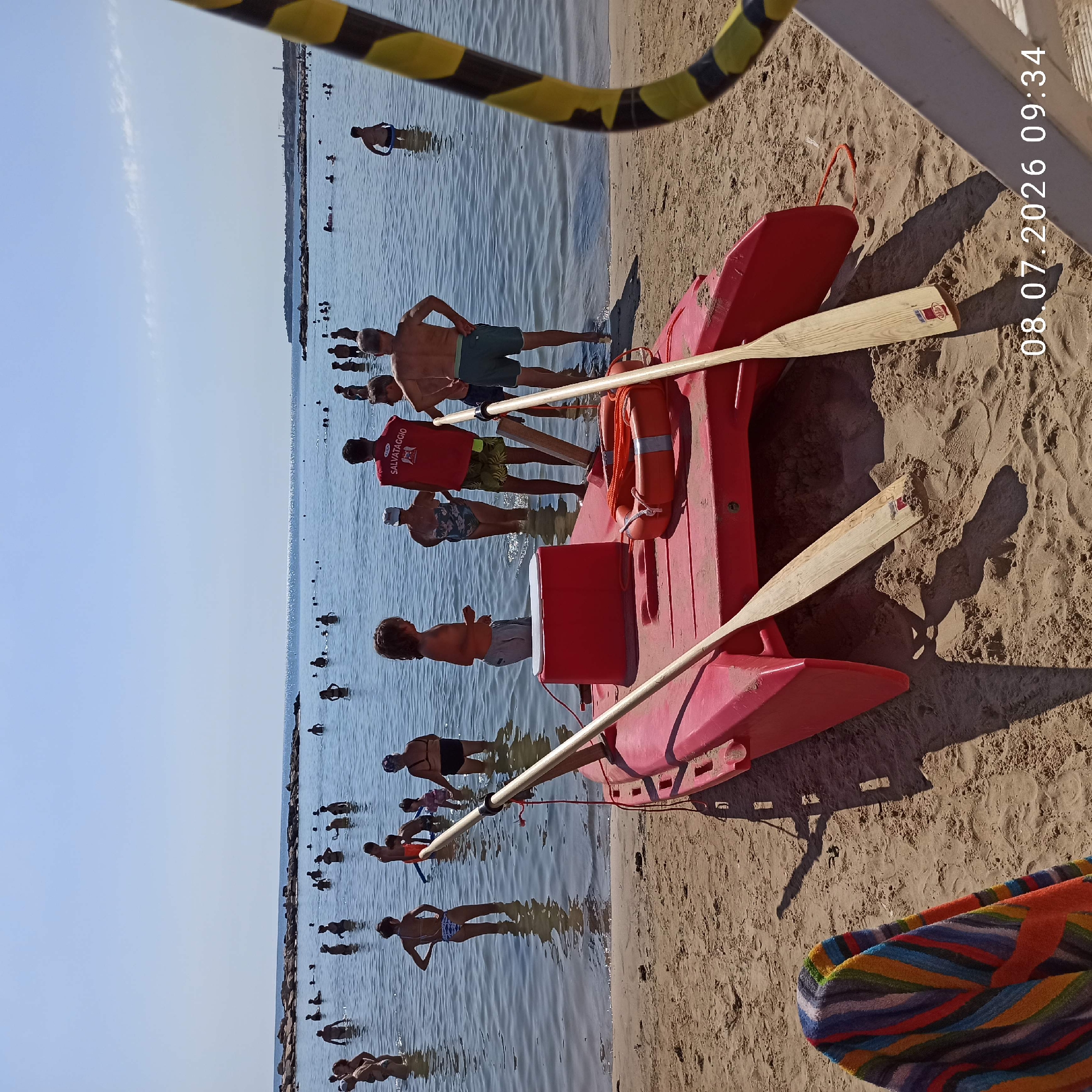 This image depicts a sunny day at a crowded beach in Trani, Italy. In the foreground, a red, inflatable rescue boat with two wooden oars is pulled up onto the sandy shore. A colorful striped towel is draped over the edge of the boat. Several people, both adults and children, are visible wading and swimming in the calm, blue water. Some individuals are standing further out in the sea. A person wearing a red vest with "SALVATAGGIO" (Italian for "Rescue") printed on it is standing near the boat, possibly a lifeguard. The background shows a clear blue sky and a rocky coastline. The overall scene is one of a typical summer day at the beach.