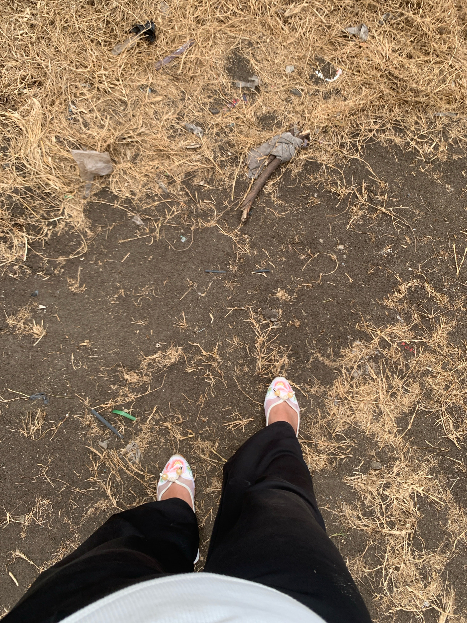 The image shows a first-person perspective of someone standing on a patch of dry grass and dirt. The ground is littered with debris, including plastic wrappers, a piece of cloth wrapped around a stick, and various small, unidentifiable objects. The person is wearing black pants and white flat shoes with a floral pattern. The location is identified as Kelurahan Tegalkamulyan, Cilacap, Indonesia.