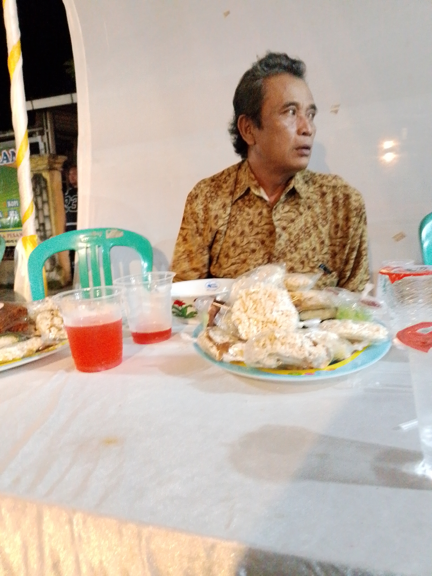 A man with graying hair and a patterned brown shirt is seated at a table covered with a white tablecloth. He is looking to his right. In front of him on the table are several plastic cups, some containing a red liquid, and a plate of food wrapped in plastic. A green plastic chair is visible behind him. The scene appears to be indoors or under a covered outdoor area, with a white background and some partially visible decorations or banners to the left, one of which has text that is partially obscured. The overall impression is of a casual gathering or meal. Based on visual cues such as the man's attire and the food items, this scene likely depicts a local event or gathering in Trangkil, Indonesia.