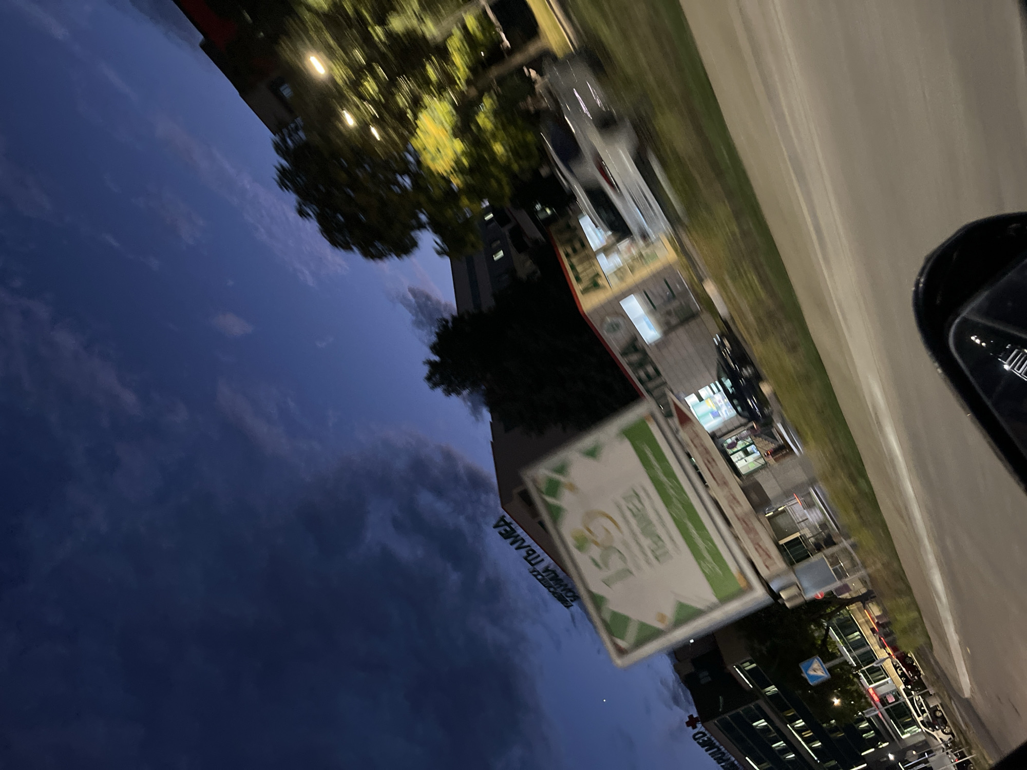The image depicts an outdoor scene at dusk, possibly in Plovdiv, Bulgaria, as suggested by the location context. The perspective is from a moving vehicle, indicated by the blur and the reflection of the car's interior in what appears to be a side mirror or window.
In the foreground, a large, rectangular sign with a green border and a white interior is prominently displayed. The sign has text and graphics, including the number "150" in gold and a shamrock logo. This sign might be advertising an event or anniversary.
Behind the sign, a building with a light-colored facade and dark-tinted windows is visible. Trees with green foliage are situated in front of the building, and streetlights with a warm glow are illuminating the area. The sky above is a deep blue, transitioning to a darker hue, with some clouds adding texture.
Further in the background, more buildings and what appear to be parked cars are discernible. One building has visible text that includes "АПТЕКА" (Apteka, meaning Pharmacy in Bulgarian) and "ПЛОВДИВ" (Plovdiv). Another building has a red cross symbol and text that reads "MED+". The overall impression is of a street or commercial area at twilight, possibly near a pharmacy or medical facility. The lighting suggests it is either early evening or late dusk. The weather appears calm.