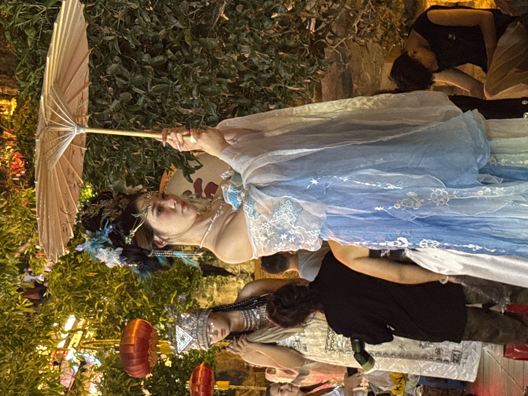 The image depicts a nighttime scene, likely in Yuzhong District, China, illuminated by ambient lighting from trees, lanterns, and possibly streetlights. In the foreground, a young woman dressed in a traditional Chinese-inspired blue and white gown with intricate embroidery and ornate headwear stands holding a light-colored parasol. Her hair is elaborately styled with blue feathers and beads. To her left, another person in similar traditional attire, also wearing a jeweled headdress, stands with hands clasped. Further to the left, a red lantern hangs amongst lush green foliage. In the middle ground, several other individuals are visible, some in traditional clothing and others in casual wear. A person in a black shirt and camouflage pants is seen from behind, partially obscured. To the right, a young man in a black sleeveless shirt is crouched down, looking towards the ground. The background consists of dense green trees, suggesting an outdoor setting, possibly a park or a traditional street market.