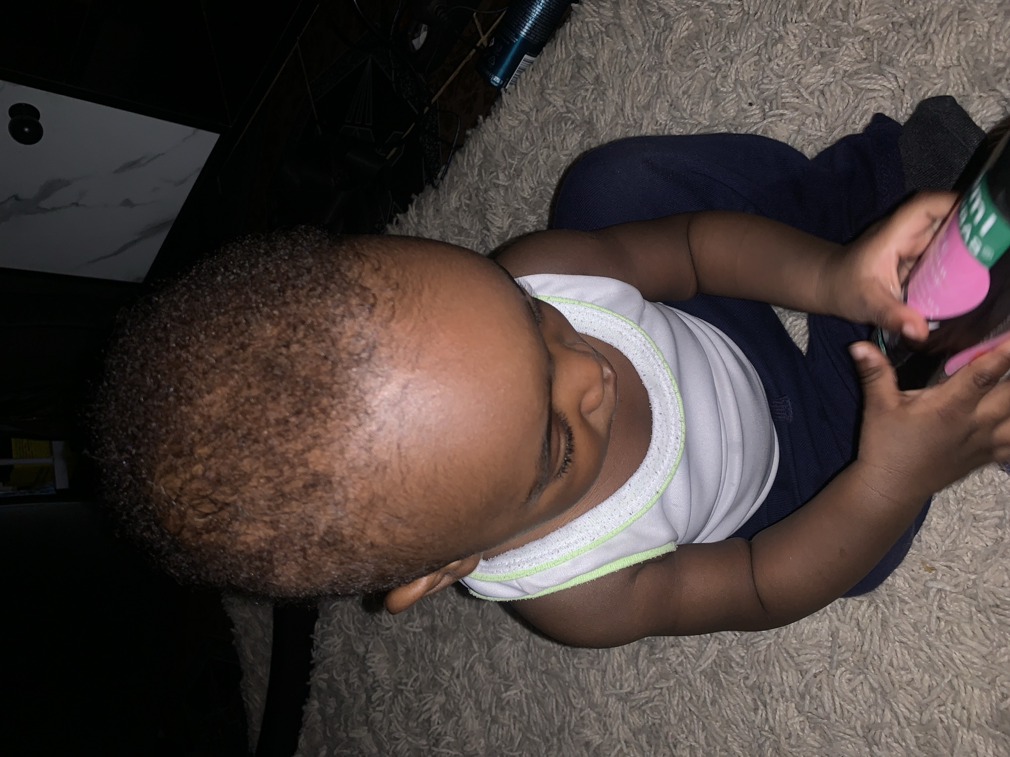 A dimly lit indoor scene shows a young Black child sitting on a cream-colored shag rug. The child is wearing a white tank top with a lime green trim and dark blue pants with dark gray socks. The child is holding a dark colored bottle with a pink and green label. The child is looking down at the bottle. The background is dark, with a portion of a marble-patterned wall and what appears to be a black piece of furniture visible. The location is stated to be Barakin Akawo, Nigeria.