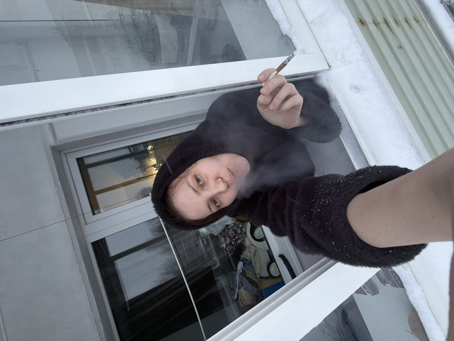 A person wearing a black, hooded furry coat stands beside a window and smokes a cigarette. Smoke plumes from their mouth. The surrounding area is snowy, particularly on the window frame and nearby surfaces. The interior reflected in the window shows a cluttered room. The image is taken from a low angle, emphasizing the person's arm and the perspective of looking up towards them.
