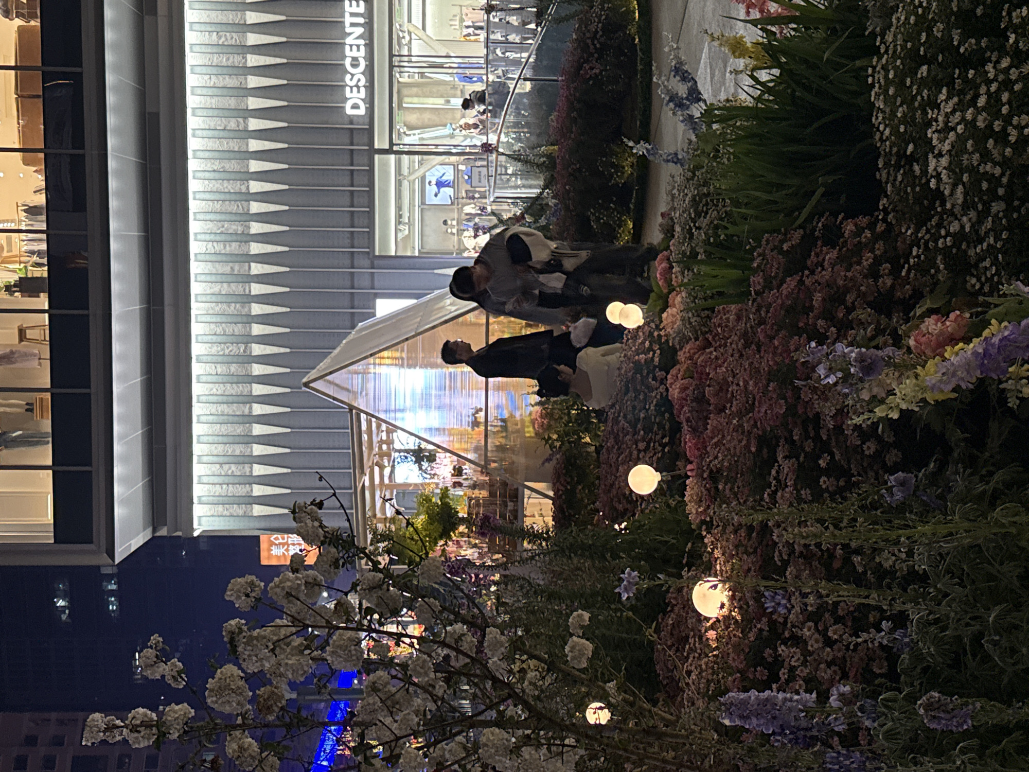 The scene depicts a nighttime view of an outdoor public space in Liangjiang, China. The foreground is occupied by meticulously arranged flower beds containing various species, including small white, pink, purple, and yellow blossoms. Several spherical white lights on slender poles are embedded within these flower beds, emitting a soft glow. A large bush with clusters of white flowers is prominent on the left side of the frame.
In the mid-ground, a translucent, illuminated pyramid-shaped structure stands. Adjacent to it, a group of four individuals is present. One individual, a female wearing a light-colored top, is seated on the ground with her back partially to the camera. Standing near the pyramid are three others: a male in a dark jacket facing left, a male in a light-grey jacket holding a mobile phone and a white garment, and a female with dark hair positioned between the two men.
The background features a multi-story modern building with large, illuminated windows displaying merchandise. The word "DESCENTE" is visible as a lit sign on the building's lower facade. Higher up, a partial orange sign with the Chinese characters "美仑" and "路形" is visible against the dark sky. The overall environment is an urban commercial area characterized by decorative landscaping and architectural lighting.