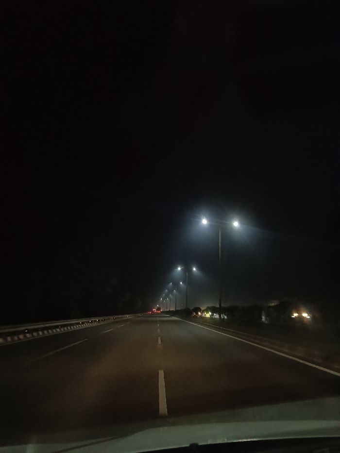 The image depicts a nighttime scene from the perspective of a vehicle traveling on a multi-lane highway. The road stretches straight ahead, illuminated by a series of bright, modern streetlights positioned along the right side. These lights, mounted on tall poles, emit a strong white glow that cuts through the darkness, creating a slight halo effect, possibly due to a thin atmospheric haze or mist.
The asphalt road is clearly marked with white dashed lines indicating lanes and solid white lines at the edges. A metal guardrail runs parallel to the left side of the highway. In the far distance, the red taillights and white headlights of other vehicles are faintly visible, suggesting the presence of moderate traffic. The top edge of what appears to be a car's dashboard or windshield is visible in the bottom foreground, indicating the image was captured from inside a moving vehicle.
The time of day is clearly night, with the sky appearing completely dark. The weather seems generally clear, though the diffusion of light from the streetlights hints at minimal atmospheric conditions. The scene evokes a sense of quiet journey or commute on a well-maintained thoroughfare. Based on the provided context, the location is likely near Umari, India. No people are discernible in the frame, and there is no visible text.