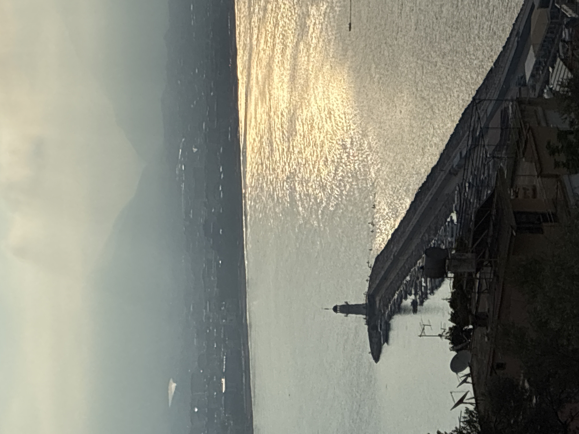 The image shows a view of the Alanya harbor in Alanya, Turkey, from an elevated perspective. A lighthouse stands at the end of a long pier, which juts out into the water. Numerous boats are docked along the sides of the pier. The water is reflective, catching the sunlight. In the foreground, the rooftops of buildings with satellite dishes and some vegetation are visible. In the background, a city skyline can be seen, with hills and mountains shrouded in haze. A small boat is visible in the distance on the water.