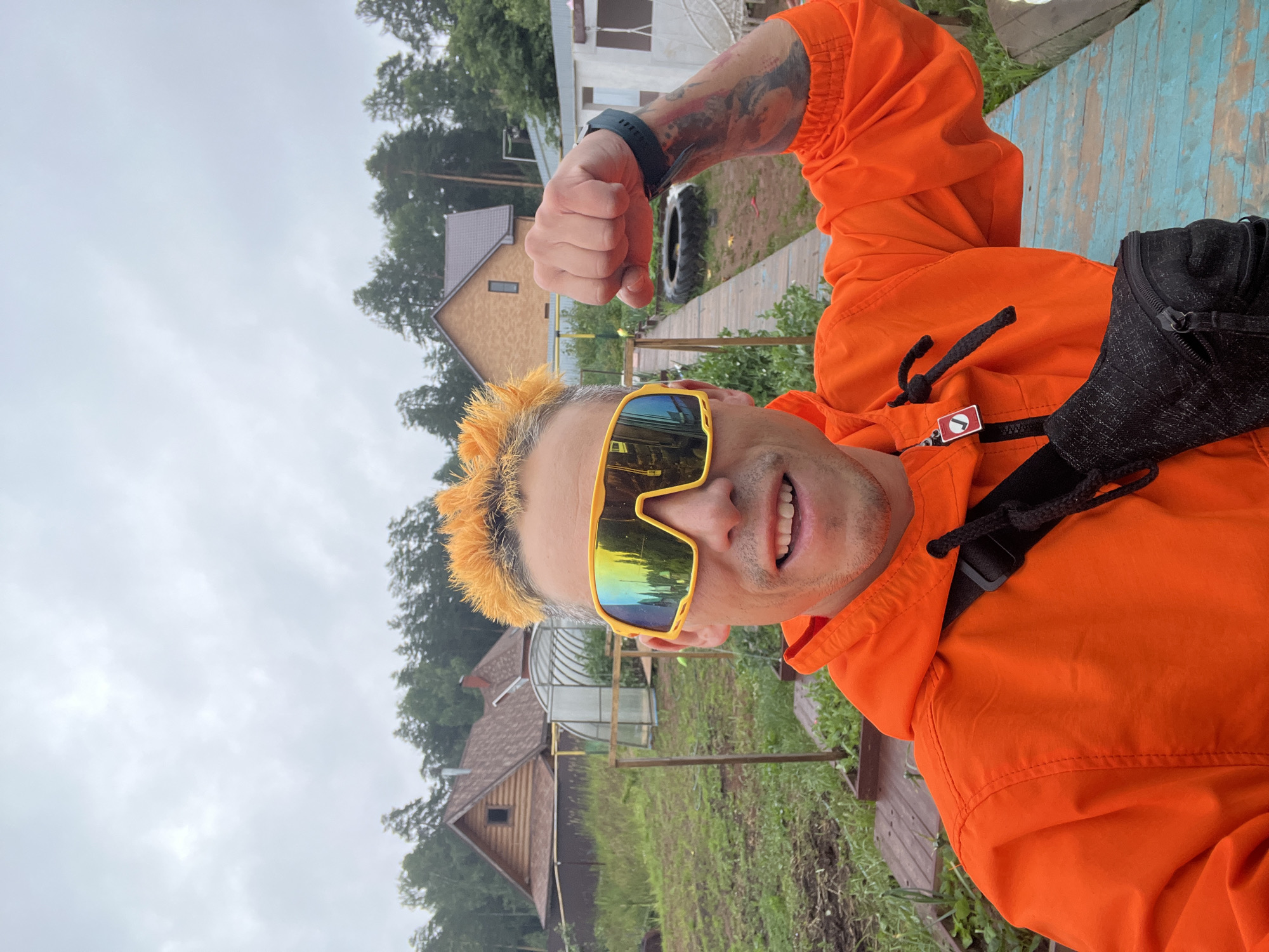A man with bright orange spiky hair and yellow mirrored sunglasses poses for a selfie outdoors. He is wearing an orange hooded jacket and a black fanny pack. His left arm is flexed, showing a tattooed sleeve. He is smiling widely.
The background suggests a rural or semi-rural setting, possibly a dacha or country house area in Chaikovsky, Russia. There are wooden buildings with brown roofs, a greenhouse, trees, and a wooden deck with blue and brown paint. The ground is covered with grass and some areas of tilled soil. The sky is overcast, indicating cloudy weather. The time of day is likely daytime due to the ambient light.
The overall mood appears playful and energetic, as suggested by the man's expression and pose, and the bright colors of his clothing and accessories. There is no visible text in the image.