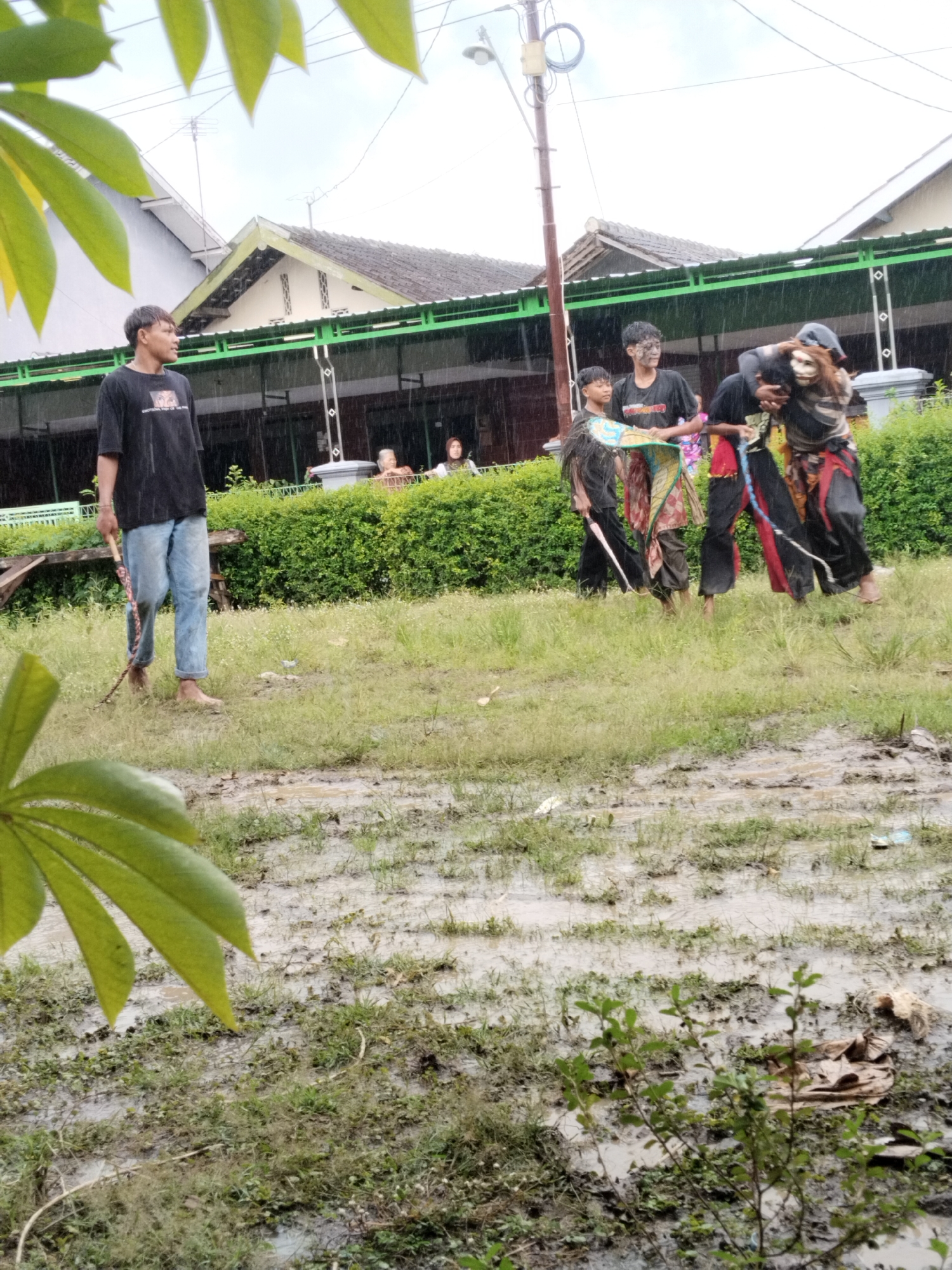 The image shows an outdoor scene in Karang Tengah, Indonesia, during a rain shower. A young man stands to the left, holding a stick, wearing a black t-shirt and blue jeans, appearing barefoot. Four other individuals are positioned towards the center of the image, seemingly involved in some kind of performance or traditional activity. One person carries another on their back who wears a mask. The two other individuals are walking alongside them, carrying decorations. The ground is wet and muddy, with scattered grass and foliage. In the background are houses.