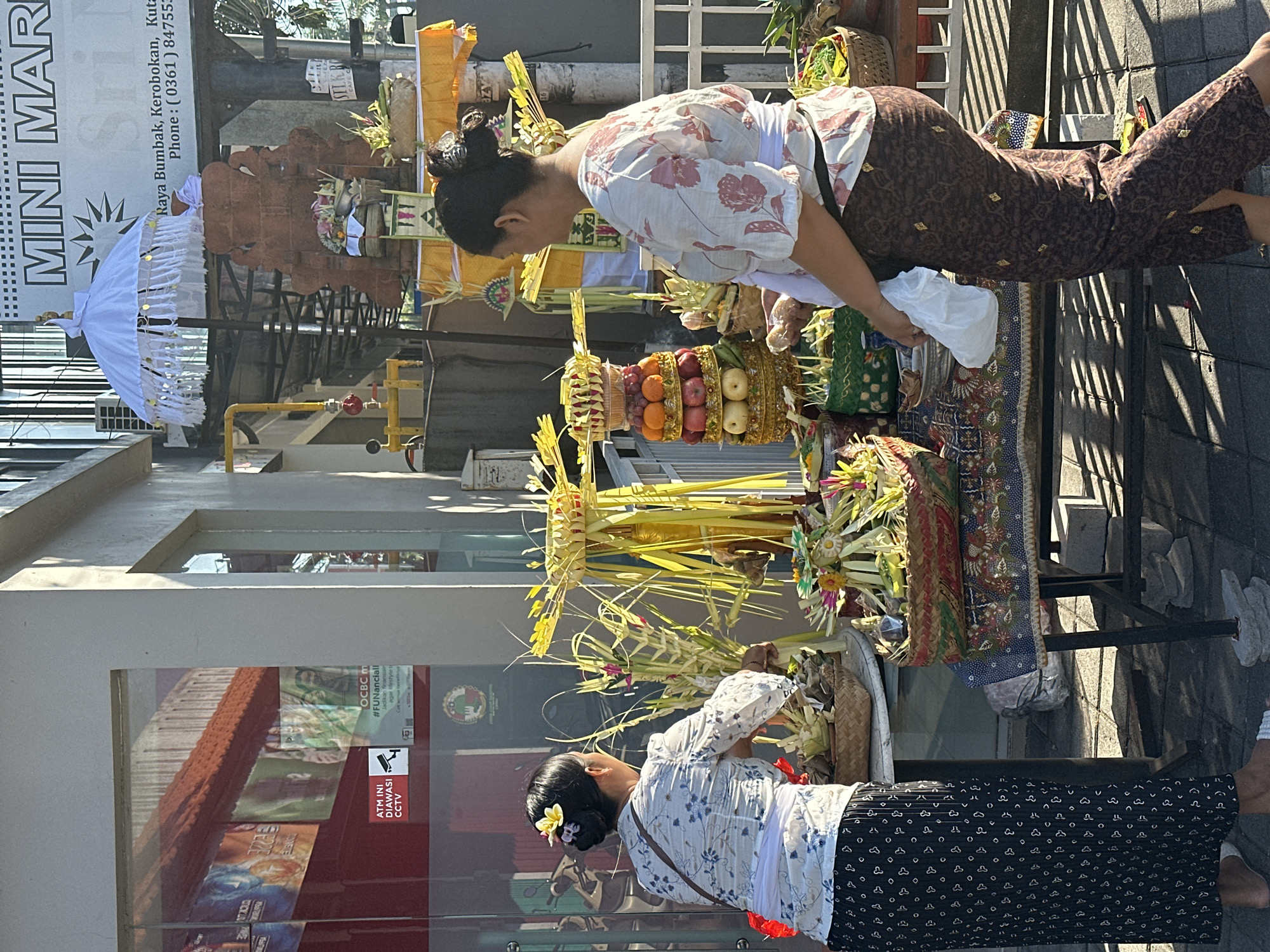 The image depicts a street scene in North Kuta, Indonesia, with two women arranging offerings outside a mini-market. The woman on the left, with a yellow flower in her hair, wears a white and blue floral top and a black patterned skirt. She is facing away from the camera, tending to offerings on a table. The woman on the right, seen from the side, wears a white floral top and a brown patterned sarong. She is holding a white plastic bag and appears to be walking or adjusting something on the table.
The offerings are elaborate and colorful, featuring woven baskets filled with fruits, flowers, and palm fronds, arranged on tables covered with patterned cloths. Behind the offerings, a brick structure with a white ceremonial umbrella stands prominently. A sign above indicates "MINI MAR" with an address and phone number below it. The mini-market's storefront is visible on the left, with red accents and banners displaying advertisements. The environment is sunny, casting shadows on the tiled ground. Additional details include an ATM with a CCTV sign inside the storefront, a gas pipe, and a white metal railing.