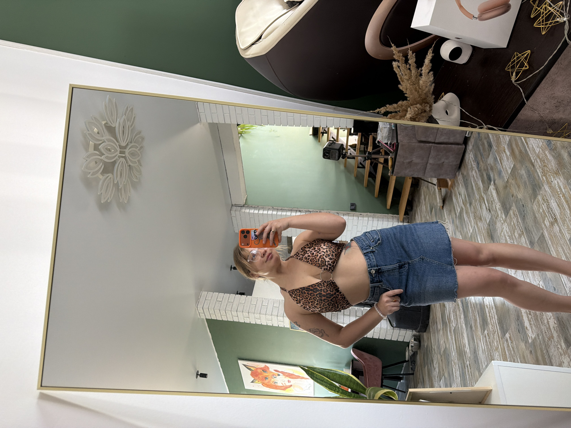 A mirror selfie shows a woman with blonde hair wearing an animal print top and denim skirt. The reflection reveals an interior with green walls, a white brick-patterned section, and a painting. The foreground includes a desk, headphones, and other decorations. It's unclear if the location is actually Большая Шильна, Russia.