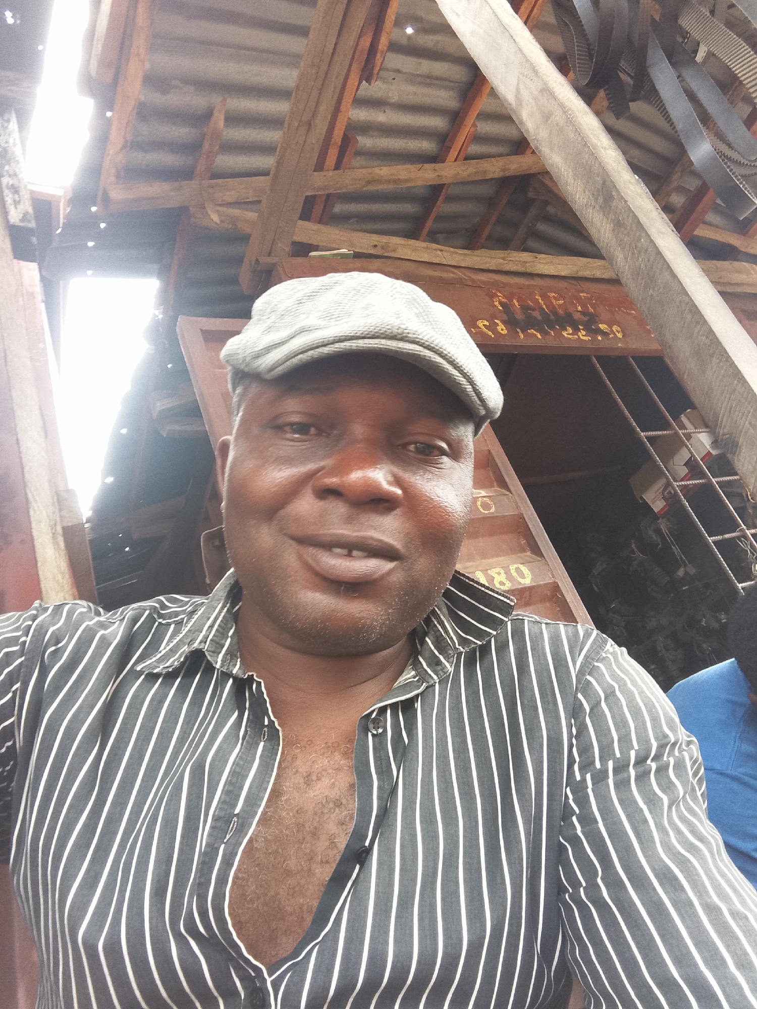A selfie taken from a low angle of a dark-skinned man with a gray newsboy cap and a gray and white striped button-down shirt. The shirt is partially unbuttoned, revealing chest hair. The background appears to be an open-air or partially enclosed structure with wooden beams and a corrugated metal roof. There are objects such as belts and other mechanical parts hanging from the structure. Writing is visible on a metal structure behind the man, possibly including numbers such as '180'. A small part of someone wearing a blue shirt is visible on the bottom right of the picture. The location is potentially Adankolo, Nigeria.