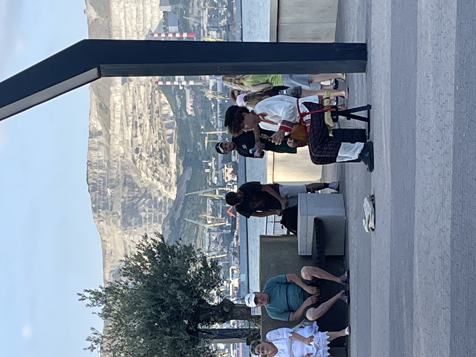 The image captures a group of people gathered outdoors, with a scenic backdrop of a rocky mountainside, a body of water, and a distant industrial port. The scene appears to be set on a paved promenade, possibly a boardwalk or pier, given the presence of a dark, angular structure overhead, suggesting a modern architectural element.
In the foreground, three individuals are seated. One person, wearing a green t-shirt and a white cap, is sitting with their legs crossed, looking towards the camera. Next to them, another person in a white outfit is partially visible, also seated. A third person, unseen, is likely sitting beside them.
Further back, a group of people are standing and interacting. One individual, dressed in a white shirt with red trim and patterned trousers, is seated on a small stool, holding a long, thin object that could be a musical instrument or prop. Another person is leaning over them, and a few other figures are standing around, some appearing to be onlookers. The clothing suggests a casual setting, possibly a warm day.
The background reveals a vast, layered mountain range with visible striations. Below the mountain, a complex of industrial structures, including cranes and smokestacks, lines the waterfront, indicating a working port. The sky is clear and light, suggesting daytime, likely late afternoon or early evening due to the quality of light. The overall mood seems relaxed and informal, with people enjoying the view and each other's company. There is no visible text in the image. The location context is Ivanovskaya, Russia.