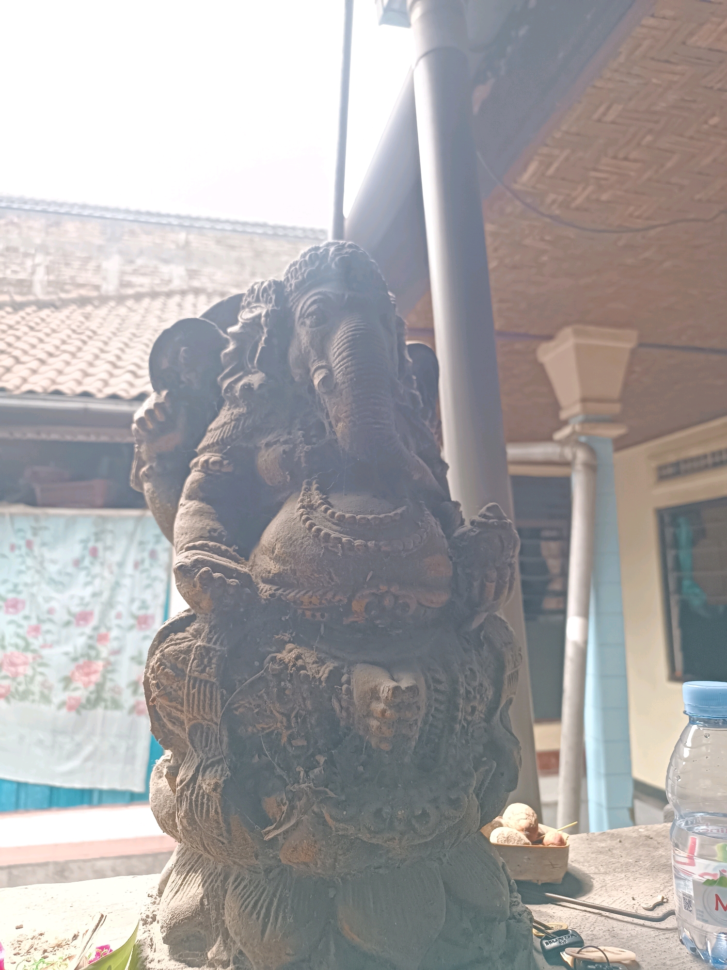 An outdoor scene captures a weathered, dark stone statue of Ganesha, the elephant-headed Hindu deity, seated on a lotus base. The statue is covered in a thin layer of dust and cobwebs, indicating it has been outdoors for some time. In the background, a tiled roof and a woven ceiling are visible, suggesting it is under some form of shelter or in a courtyard. To the right, a partially filled plastic bottle of water stands on a concrete surface, next to a small woven container holding several small, round objects, possibly food items. A few keys and a metal object are also present on the surface near the bottle. The lighting is bright, suggesting daylight, and a light-colored curtain with a floral pattern hangs behind the statue.