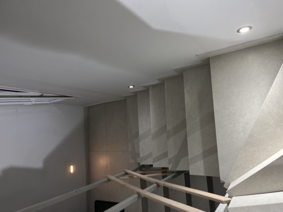 The image shows a set of stairs in a residential building in Limenaria, Greece. The stairs are made of light-colored stone and are illuminated by small, recessed light fixtures on the wall. The handrail is made of metal and wood. The floor is tiled. The scene is empty of people. The image captures the staircase from the bottom, looking up towards the top. The stairs are set against a white wall and are part of an interior space. There is a window on the right side of the image, looking out towards an unknown exterior. The image is taken at a slightly downward angle, showing the staircase from the bottom. The image is well lit and the colors are muted. The scene conveys a sense of simplicity and peace.