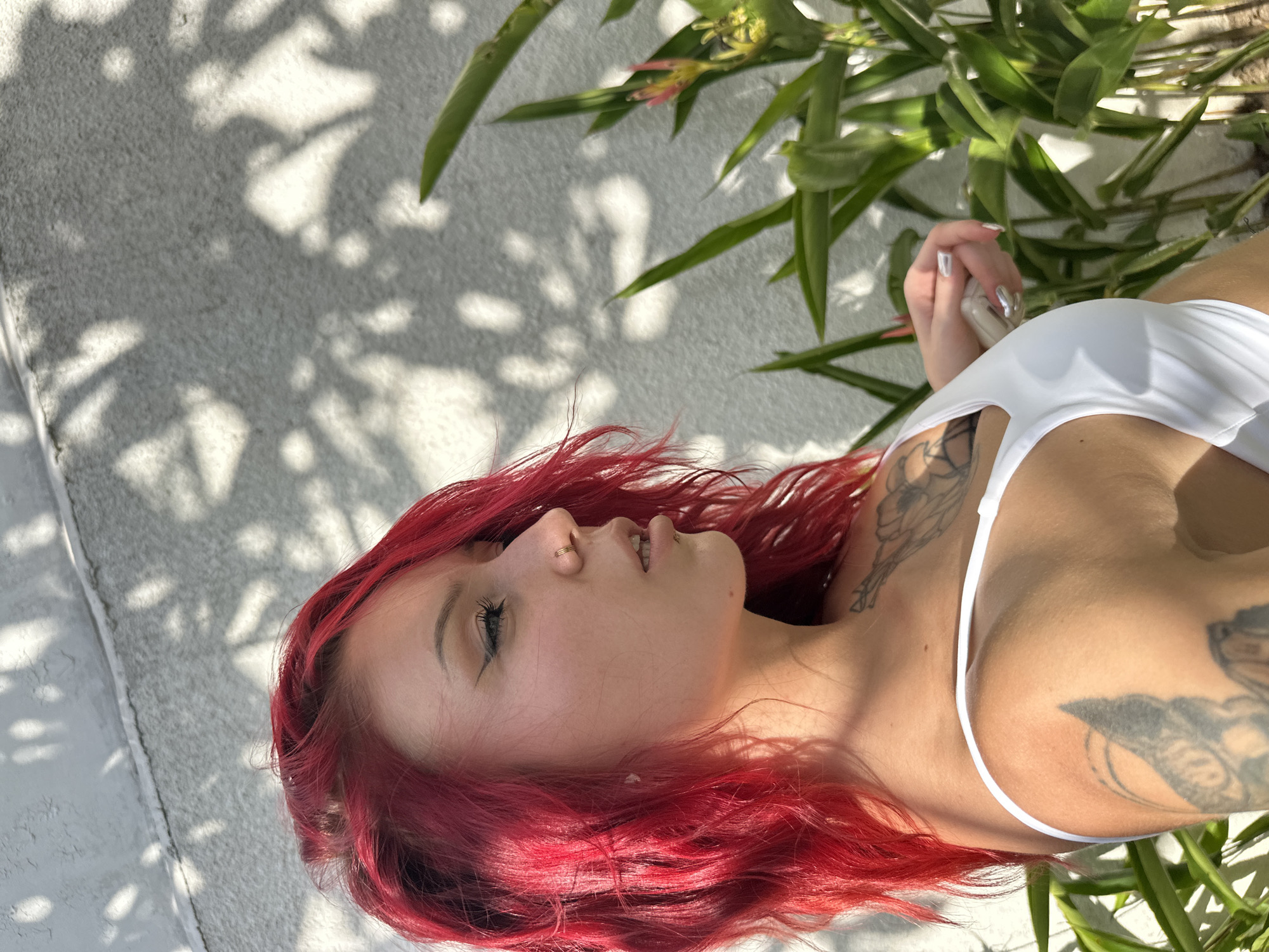 A pale-skinned female with red hair and visible tattoos is laying down outside, under an area with leafy plant growth. The background consists of a light-colored, textured wall with dappled shadows. The location is unknown; any reference to North Kuta, Indonesia would be speculative.