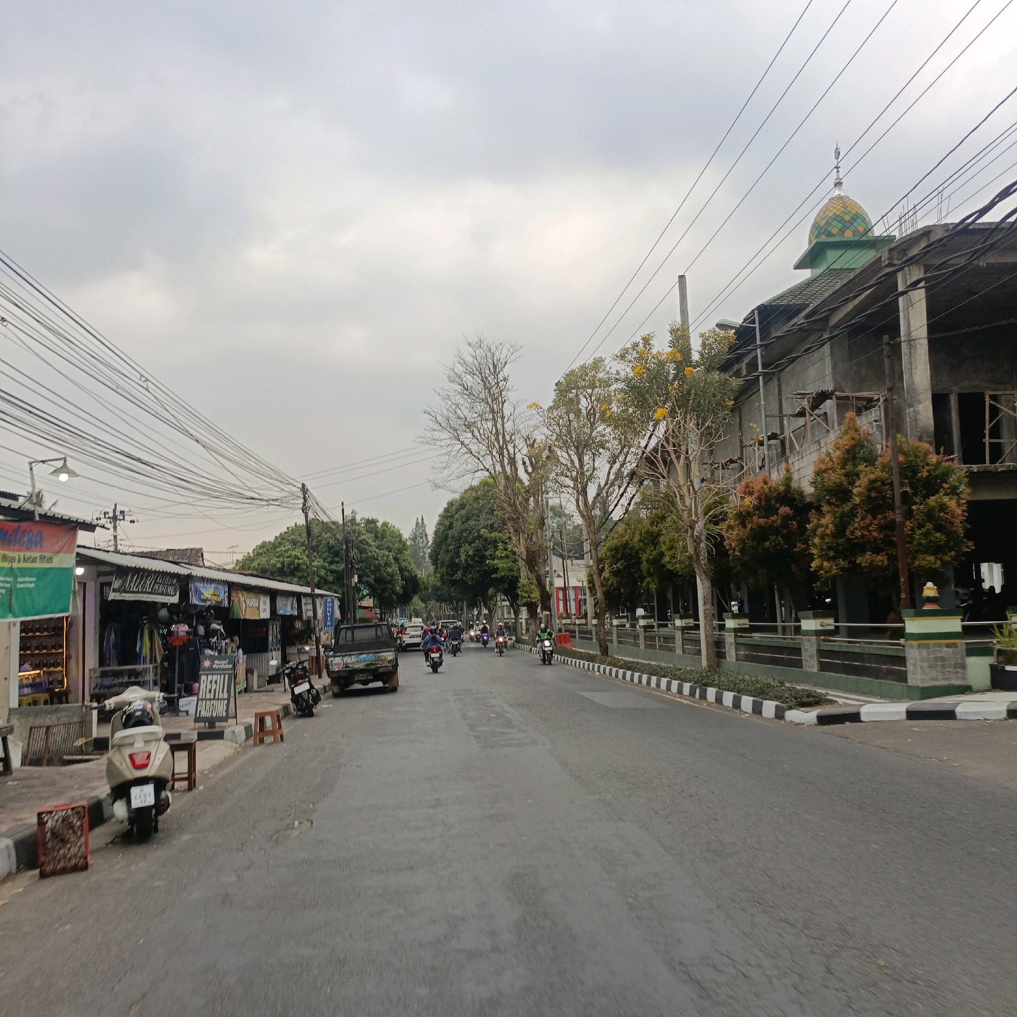 The image depicts a street scene in Salatiga, Indonesia, under an overcast sky. A road runs through the center, with traffic consisting of motorcycles and a pickup truck. On the left side of the road are several small businesses with awnings and signage. One shop displays clothing and has a sign for "Refill Parfum." A light-colored scooter is parked on the sidewalk in front of this shop, next to a small wooden stool. Further down the street on the left, a more modern building with a green roof and a decorative yellow and green dome, likely a mosque, is visible. The right side of the road features a building under construction, with scaffolding and concrete walls. Trees line the sidewalk on the right, some with yellow flowers, and a decorative fence with green and white elements separates the sidewalk from the building. Power lines crisscross the sky above the scene.