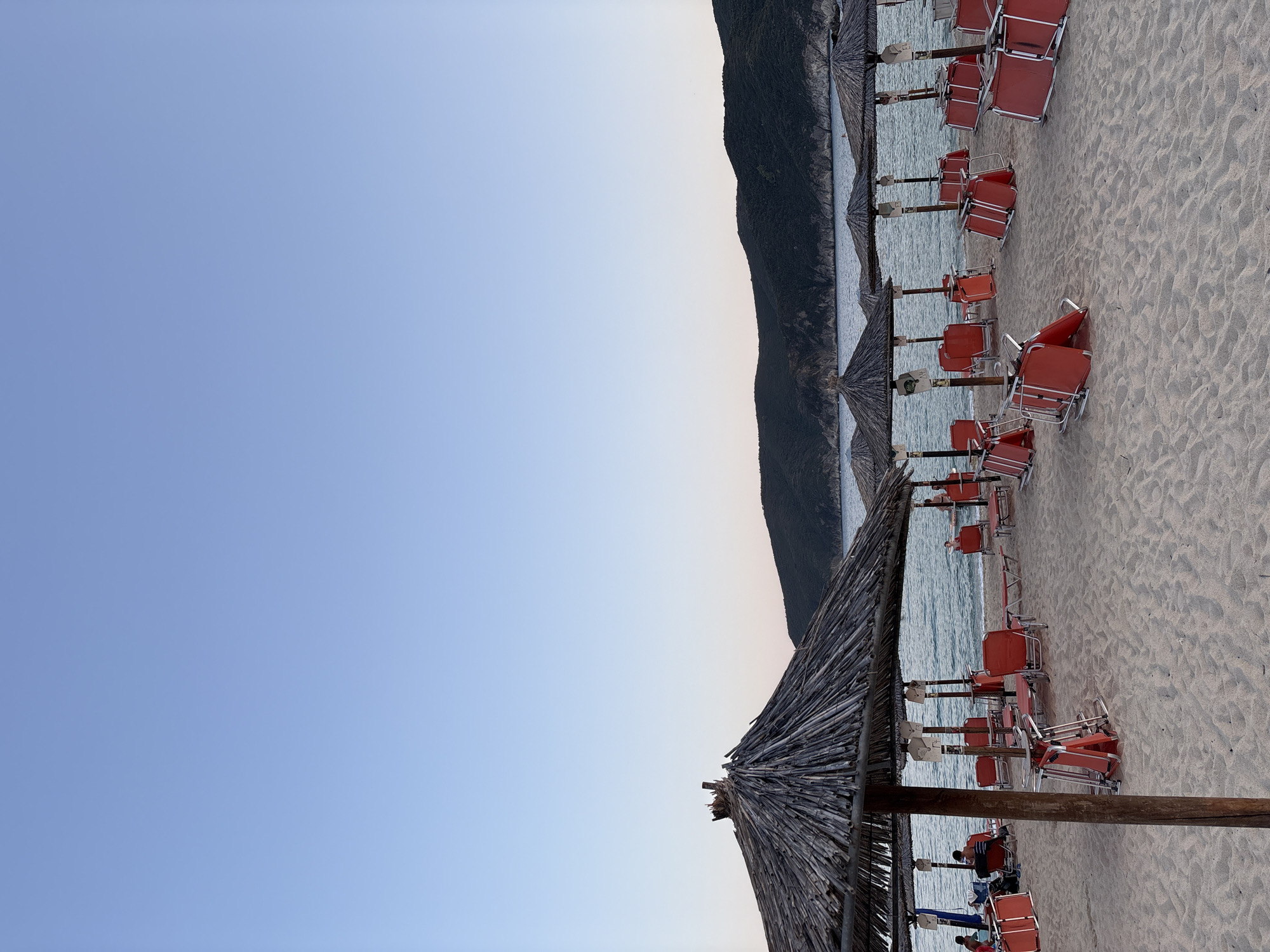 The image depicts a beach scene in Toroni Municipal Unit, Greece, likely in the late afternoon or early evening. The foreground is dominated by a sandy beach with numerous folded red lounge chairs and a few unfolded ones. Several straw-thatched umbrellas are erected on wooden poles, providing shade. In the mid-ground, the calm sea stretches towards a dark, tree-covered hillside that forms the coastline. The sky is a clear, gradient blue, transitioning from a lighter hue near the horizon to a deeper shade at the top of the frame. A few individuals are visible on the beach, some sitting in chairs under the umbrellas, while others appear to be standing near the water's edge. The overall atmosphere is peaceful and suggests the end of a day at the beach.