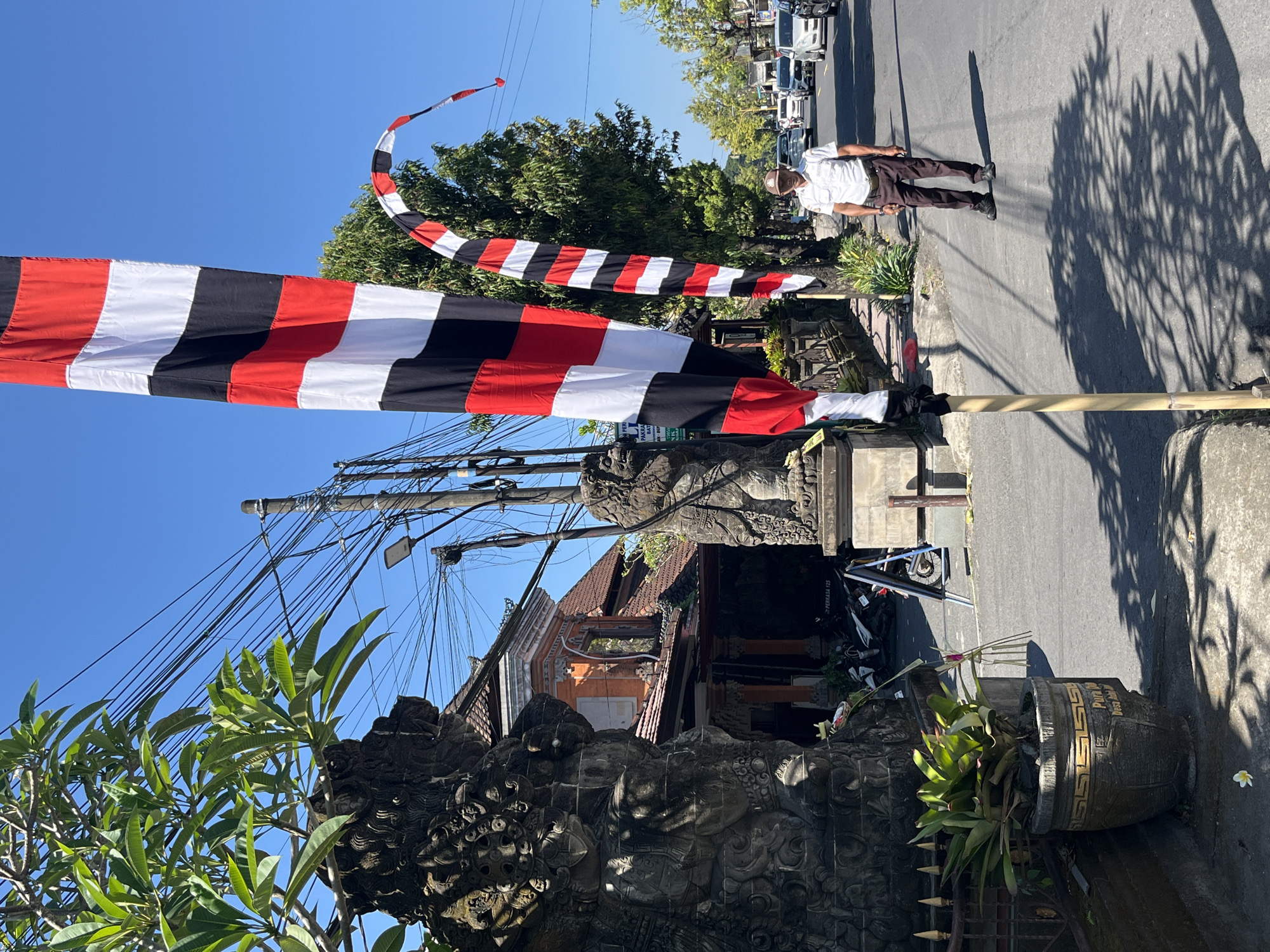 The image depicts a street scene in Sukawati, Indonesia, likely on a sunny day, given the clear blue sky and strong shadows. In the foreground, a tall, vertical flag with alternating red, white, and black stripes stands prominently. To its right, a curved banner of the same color scheme arcs upwards.
On the left side of the image, a stone statue, possibly a guardian figure, is partially visible, adorned with intricate carvings. Adjacent to it is a large potted plant with broad green leaves. Behind the statue, a building with orange walls and a tiled roof is visible, featuring traditional architectural elements. More stone sculptures and foliage line the pathway.
A paved road runs through the scene, with several vehicles, including cars and motorcycles, visible in the background, suggesting a moderately busy street. On the right side of the road, a man wearing a white short-sleeved shirt, dark trousers, and a cap stands facing forward. His shadow is cast on the asphalt. The environment is characterized by overhead power lines strung between poles, common in many urban and semi-urban areas. The overall atmosphere suggests a cultural and residential setting within the town.