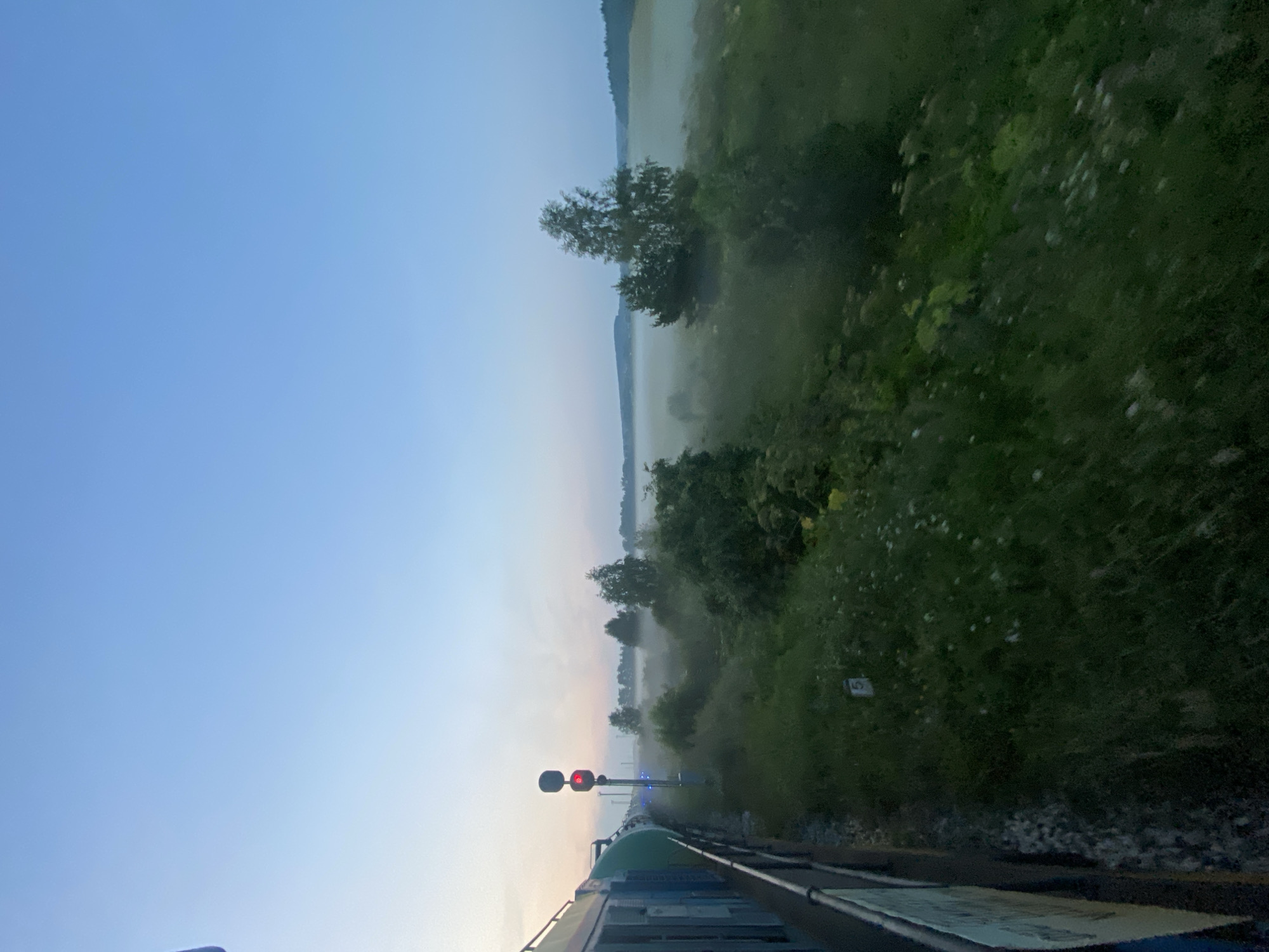 The image captures a view from a moving train on a clear, cool morning, likely near Kauchuk Stantsiya, Russia. The sky transitions from a pale yellow near the horizon to a soft blue overhead. In the foreground, the side of a green and white train is visible, along with a railing. To the right of the tracks, a single railway signal displays a red light. Beyond the signal, a misty field stretches into the distance, dotted with clusters of trees. The ground closest to the tracks is covered in lush green grass with scattered white wildflowers and a small white marker labeled "5". The overall atmosphere is serene and rural, with the mist adding a touch of mystery to the landscape.