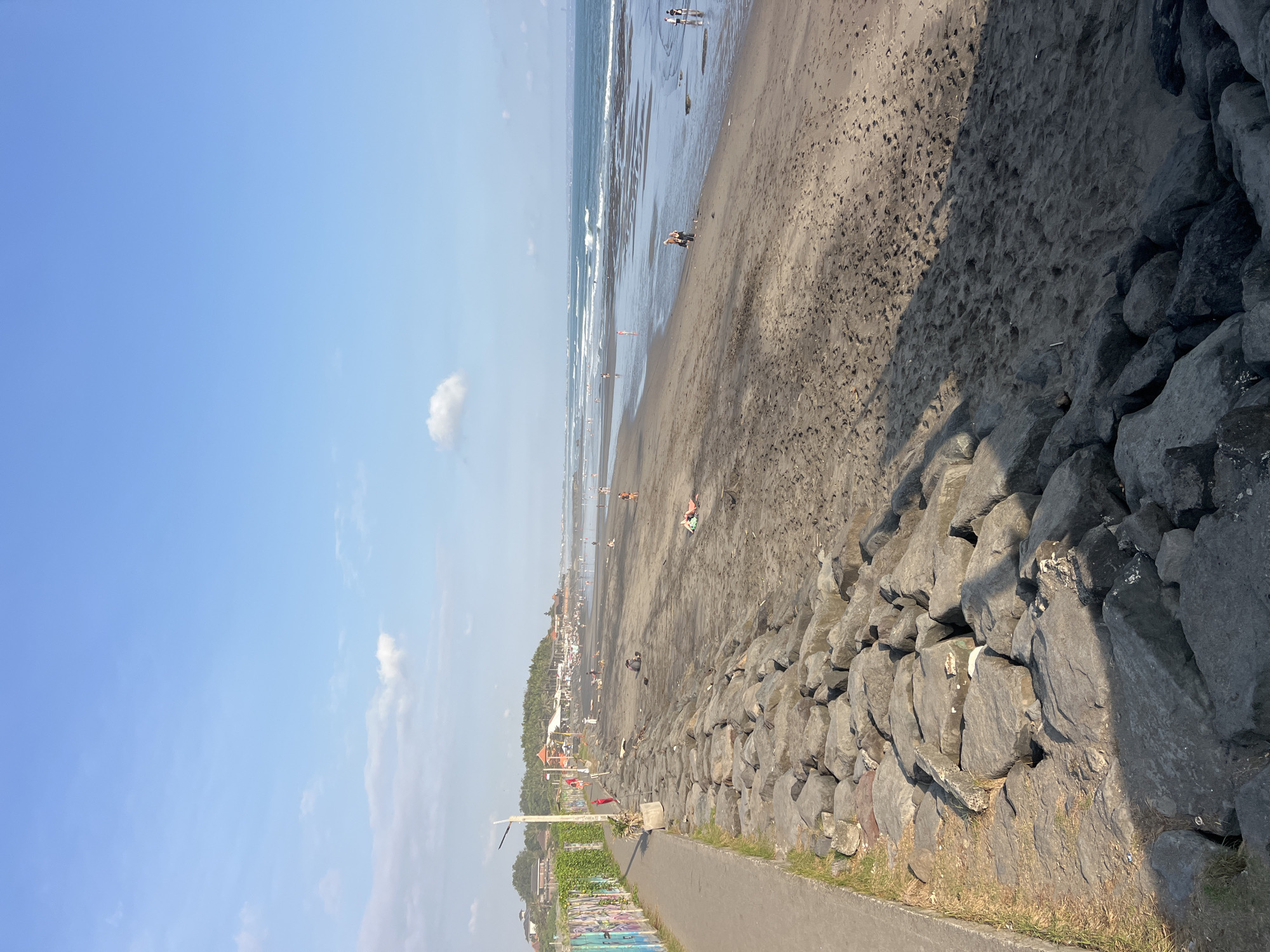 The image shows a wide, dark sand beach with a rocky embankment in the foreground. A paved walkway runs alongside the embankment, bordered by a graffiti-covered wall on the left. The ocean is visible in the background, with gentle waves and a hazy horizon. Several people are scattered across the beach, some walking, some sitting, and some lying down. In the distance, there are buildings and palm trees, suggesting a populated area. The sky is a clear blue with a few scattered white clouds. The scene appears to be from a sunny day, likely in the afternoon, given the shadows. This beach is located in Mengwi, Indonesia, an area known for its coastal attractions.