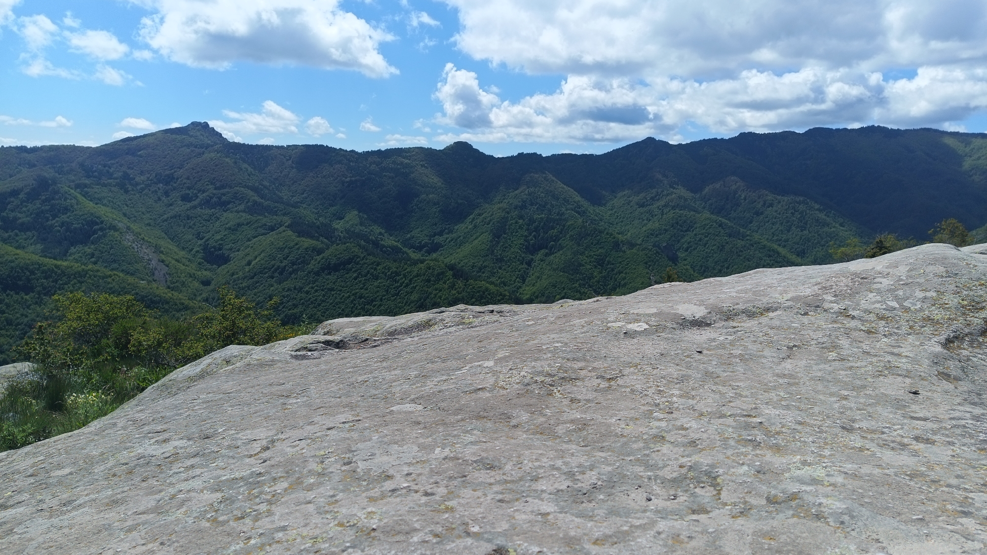 The image shows a mountainous landscape under a partly cloudy sky. The foreground features a large, flat rock surface with a rough texture. In the midground, there is a series of rolling, forested hills and mountains, covered in dense green vegetation. The sky is blue with scattered white clouds. The location is Sini Vrah, Bulgaria.