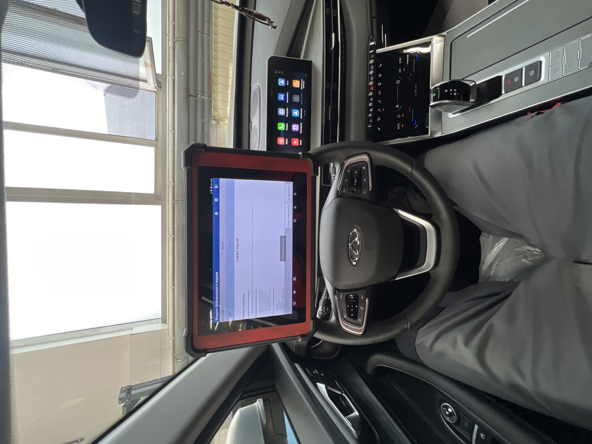 An interior view of a modern Chery vehicle, purportedly in the city of Izhevsk, Russia, reveals a diagnostic procedure underway. A person's legs, clad in grey trousers, are visible occupying the driver's seat, with clear plastic protective sheeting covering the floor beneath them. A red and black ruggedized diagnostic tablet is securely mounted on the steering wheel via an articulated arm. The tablet's screen displays Russian text, indicating "Модель Информация о версии" (Model Information about Version) and identifies the software as "CHERY V60.99." Details regarding "Software introduction" list basic functions like "Read car composite information" and "Read car fault code," along with special functions for "Engine" and "ABS/Anti-lock Brake System." An "Authenticate success" button is visible. The tablet's status bar shows 100% battery and the time 6:19.
The car's interior features a multi-function steering wheel with a "CHERY" logo and "SRS AIRBAG" label. To the right, a wide infotainment screen displays a grid of application icons, including "Android Auto," "Apple CarPlay," and "Обновление системы" (System Update). Below this, a touch-sensitive climate control panel indicates "88" degrees on both sides for dual-zone control, with buttons for "AUTO," "A/C," "SYNC," and "CLEAN AIR." An electronic gear selector is positioned in "P" (Park), flanked by buttons for the electronic parking brake and auto-hold function. The driver's side door panel is dark, featuring a silver door handle and window controls.
Through the windshield, a bright, indoor environment consistent with a service bay or garage is visible. This area includes large, multi-pane windows with light-colored frames; one window features an exterior metallic mesh screen. A high, white ceiling is also discernible. A rearview mirror and a small decorative charm are partially visible at the top right of the image.