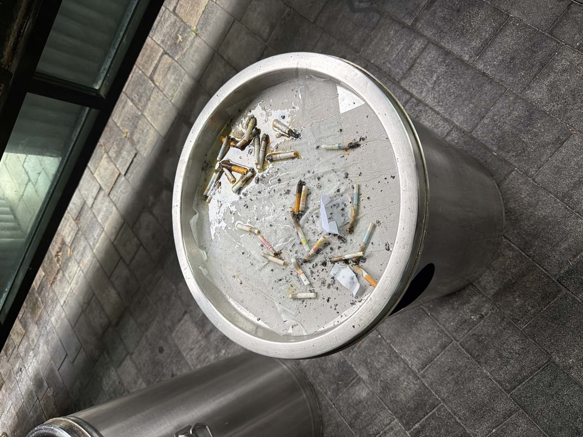 An overhead shot depicts a circular, metal ashtray filled with cigarette butts, ash, and small pieces of white paper. The ashtray sits on gray pavement, a building with a dark window is visible in the background. The location is Gimpo-si, South Korea.