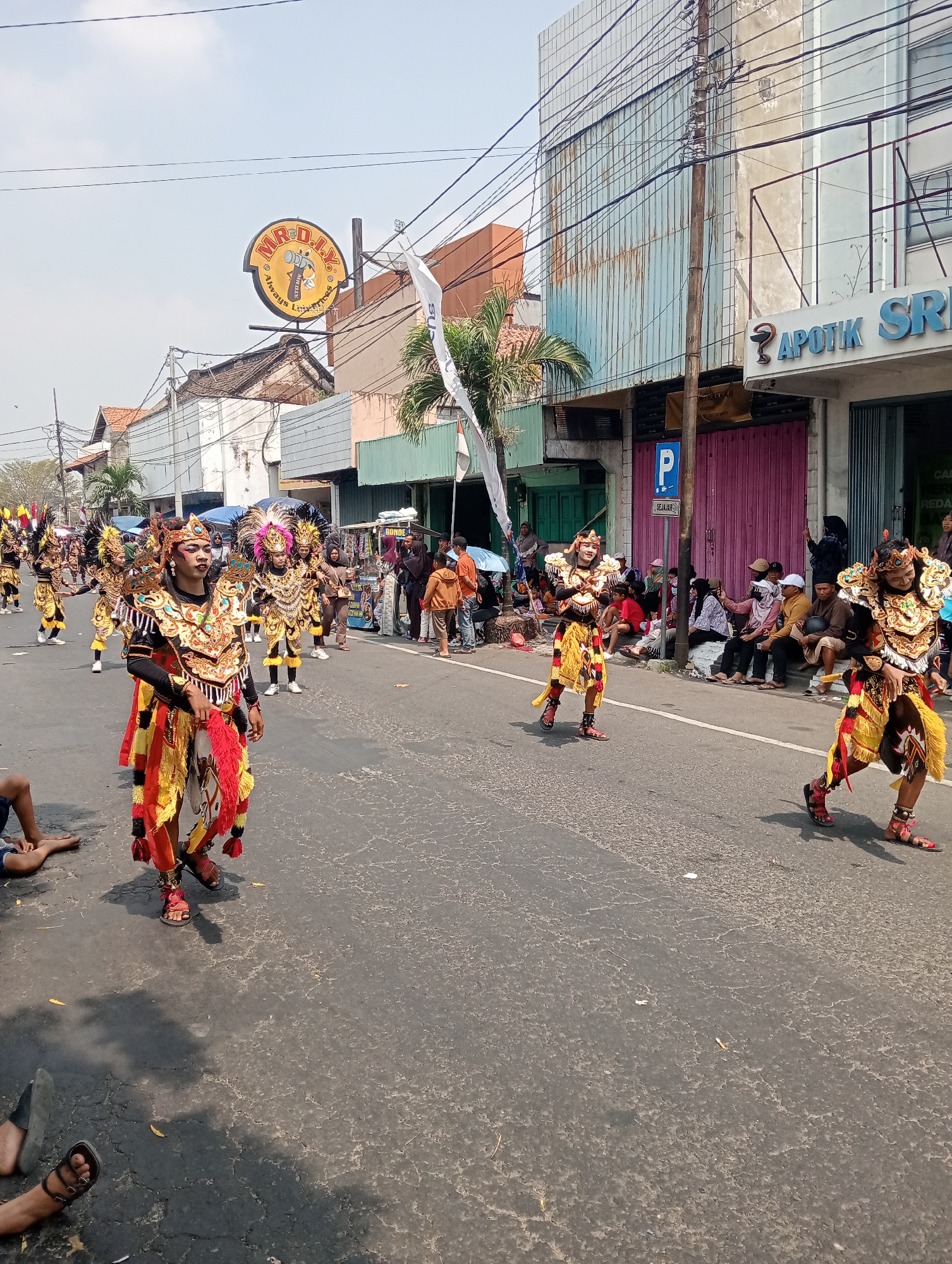 The image captures a daytime street scene in Demak, Indonesia, likely during a cultural parade or festival. In the foreground, several individuals adorned in vibrant, traditional costumes are walking down a paved street. These costumes feature elaborate headdresses with feathers, ornate gold and black chest plates, and flowing red, yellow, and black fabric skirts. Some of the performers have face paint or makeup. The street is lined with buildings on both sides, which appear to be shops and businesses. One prominent sign visible is for "MR. D.I.Y.", a retail chain. Another sign reads "APOTIK SRI". Spectators are visible along the sides of the street, some sitting on the curb and others standing. The sky is clear and bright, indicating sunny weather. The overall atmosphere suggests a lively public event.