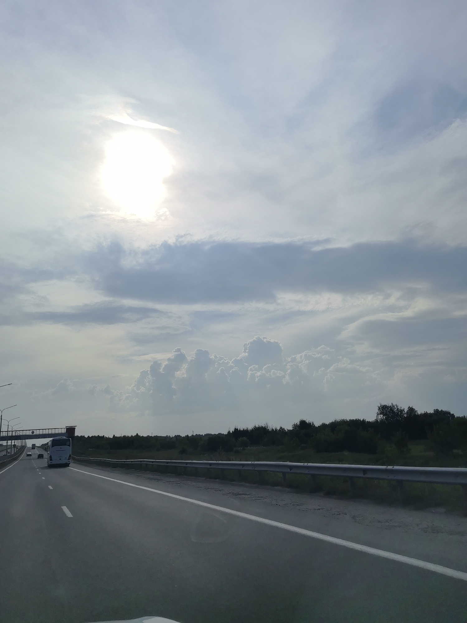 The image depicts a highway scene with a bright sun partially obscured by clouds. A white bus is visible driving on the road, along with other vehicles in the distance. The road has clear white lane markings. A metal guardrail runs along the side of the highway, separating it from a grassy area and a line of trees in the background. There are also streetlights visible on the left side of the image. The sky is partly cloudy with dramatic cloud formations. The scene appears to be set in a rural or suburban area outside of a city, possibly near Timiryazevsky, Russia, given the context.